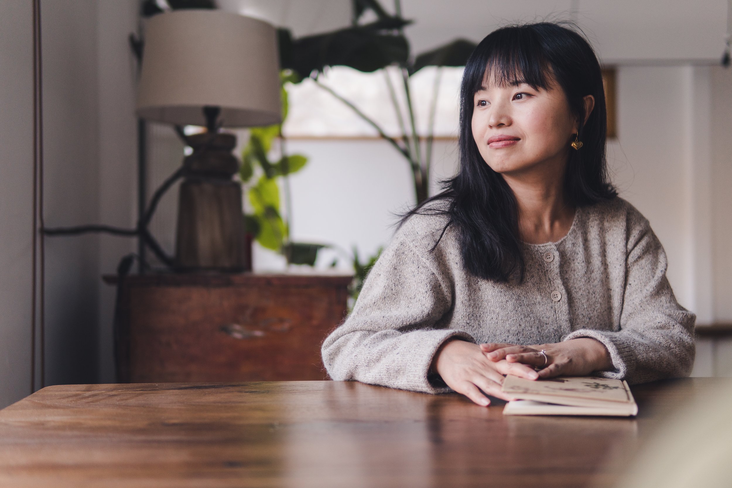 A woman with shoulder-length black hair sitting at a wooden table with an open notebook in front of her. She is wearing a beige cardigan and earrings, looking to the side with a neutral expression. There is a lamp and some green plants in the backgro