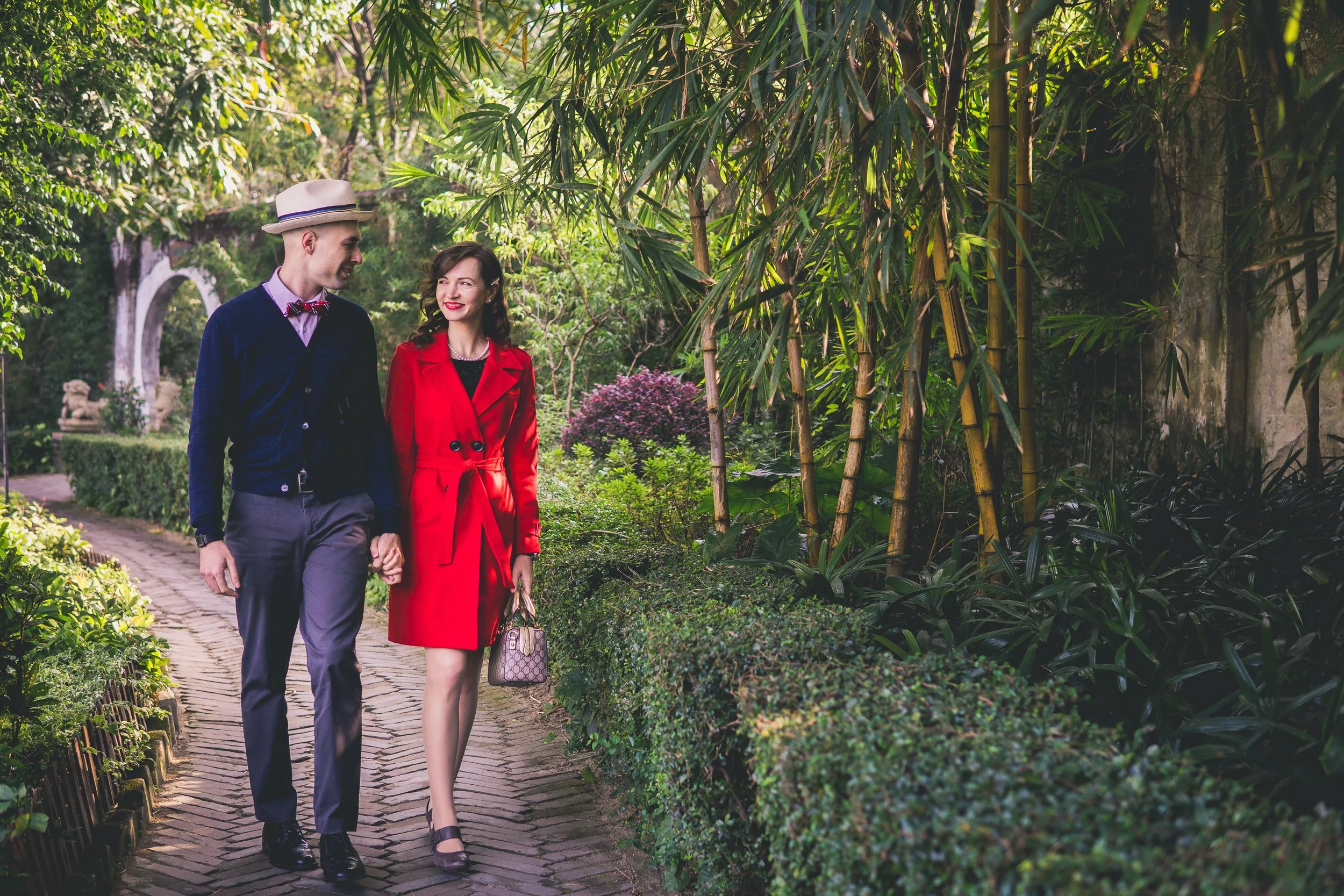 A man and woman walking hand in hand through a lush, green garden. The man is wearing a beige hat, dark blue cardigan, and gray trousers, while the woman is dressed in a red coat and carrying a purse. They are smiling at each other.