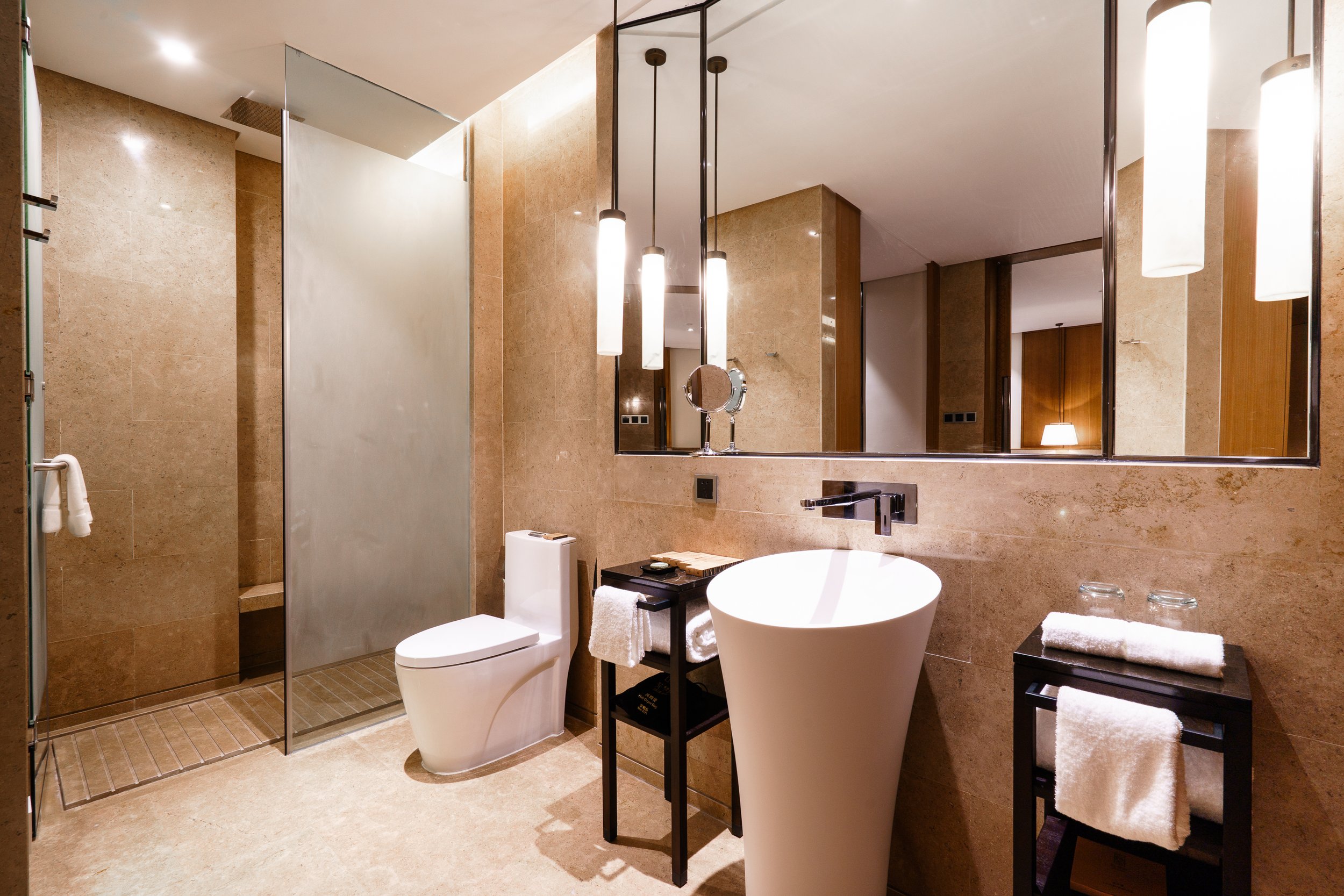 Modern bathroom with beige tile walls and floor, featuring a walk-in shower, white toilet, pedestal sink, large mirror, and black side tables with towels and glassware.