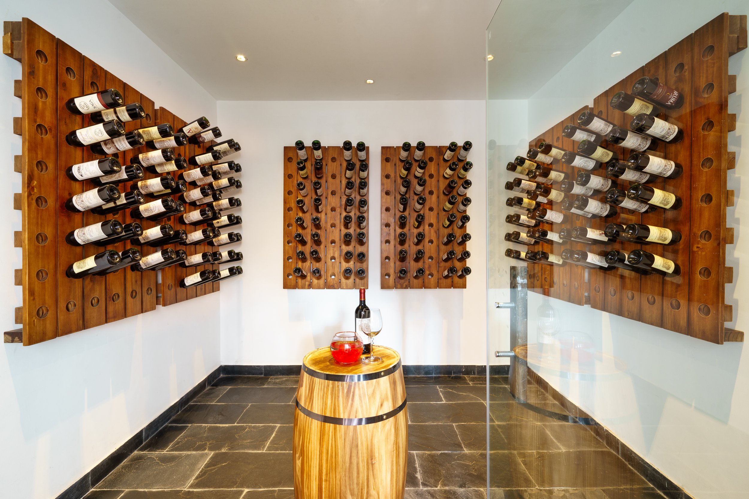 Wine bottles are stored on wooden racks on the walls of a room, with a small barrel table in the center holding a bottle of wine, a glass of red wine, and a decanter.