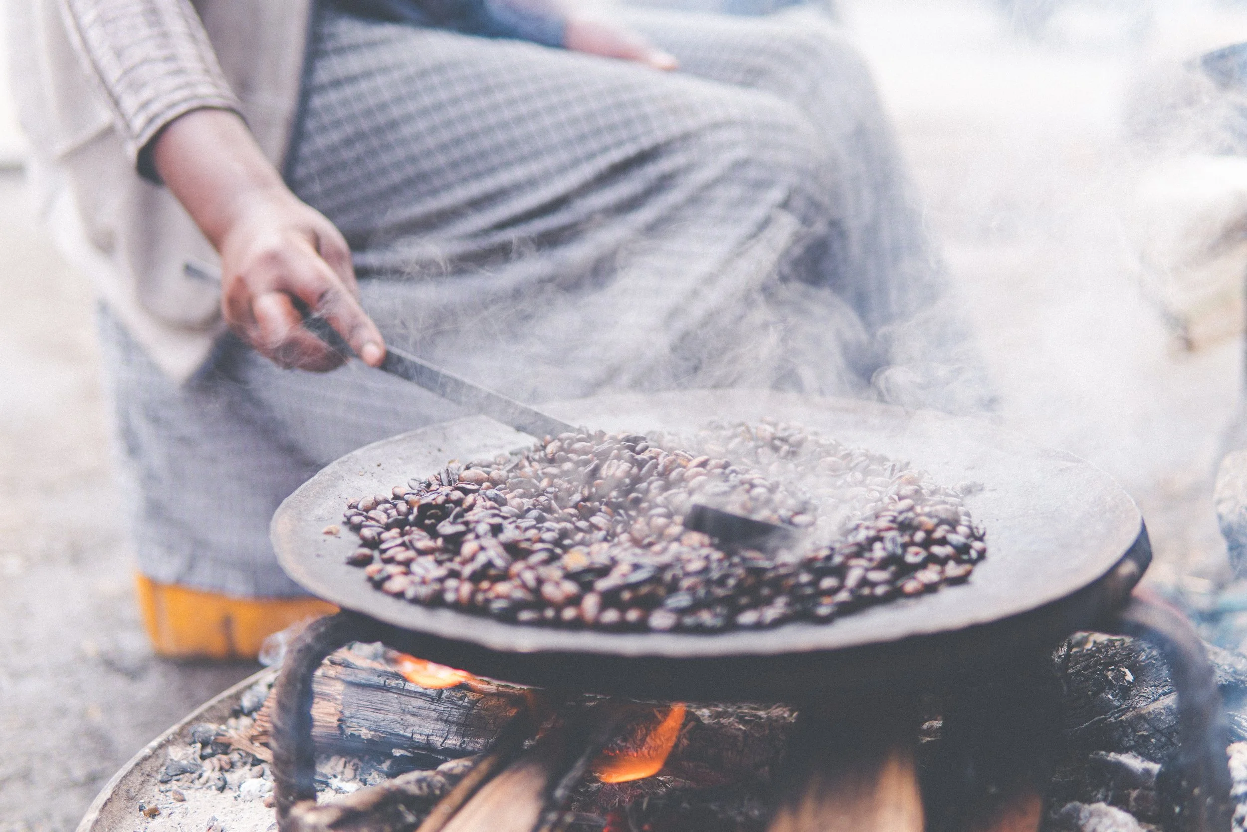 Person cooking coffee beans on an outdoor open flame stove, with steam rising from the beans.