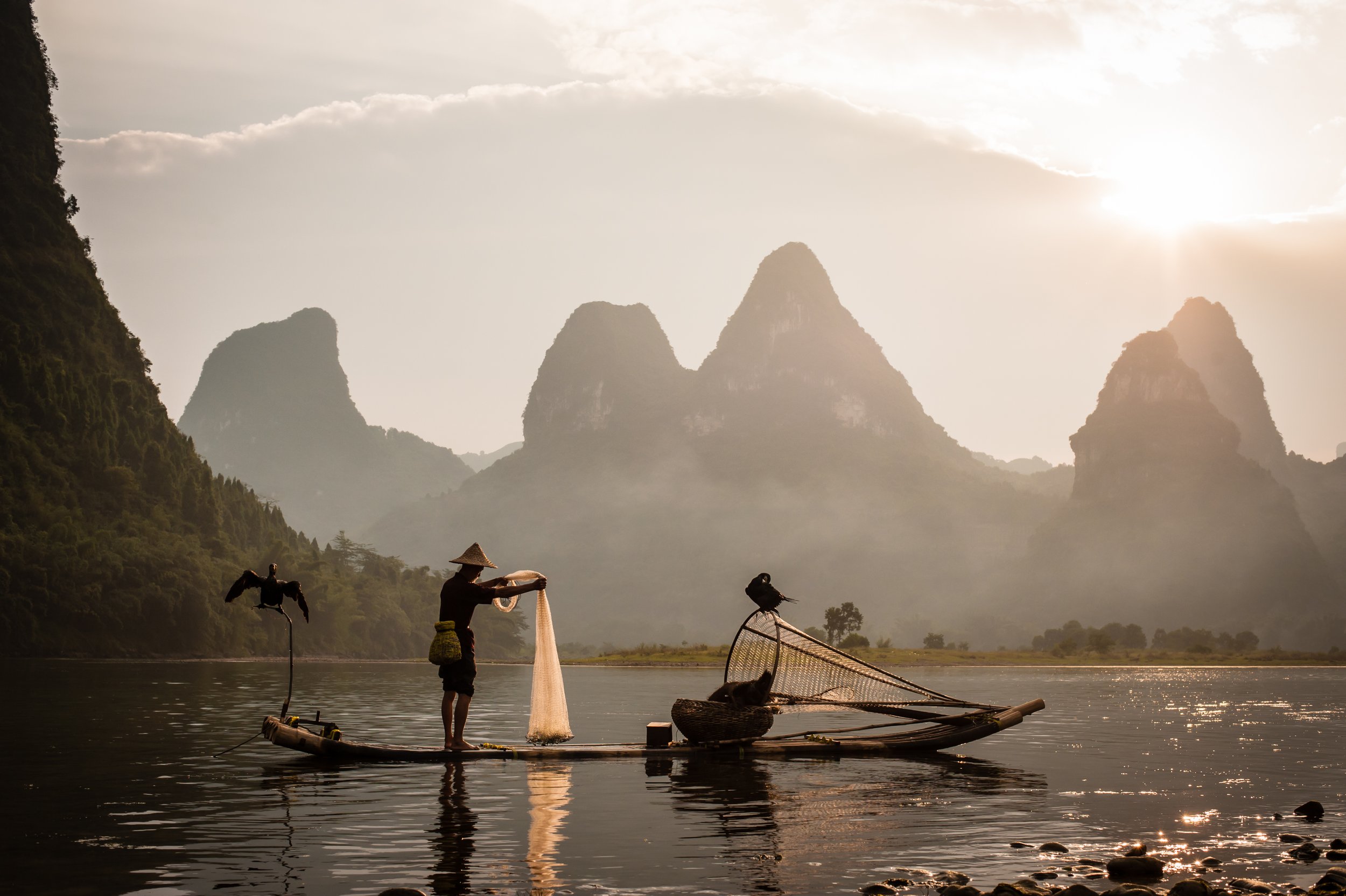 Fisherman on bamboo raft with birds, fishing net, and fish trap, surrounded by mountains and river in the background during sunset.
