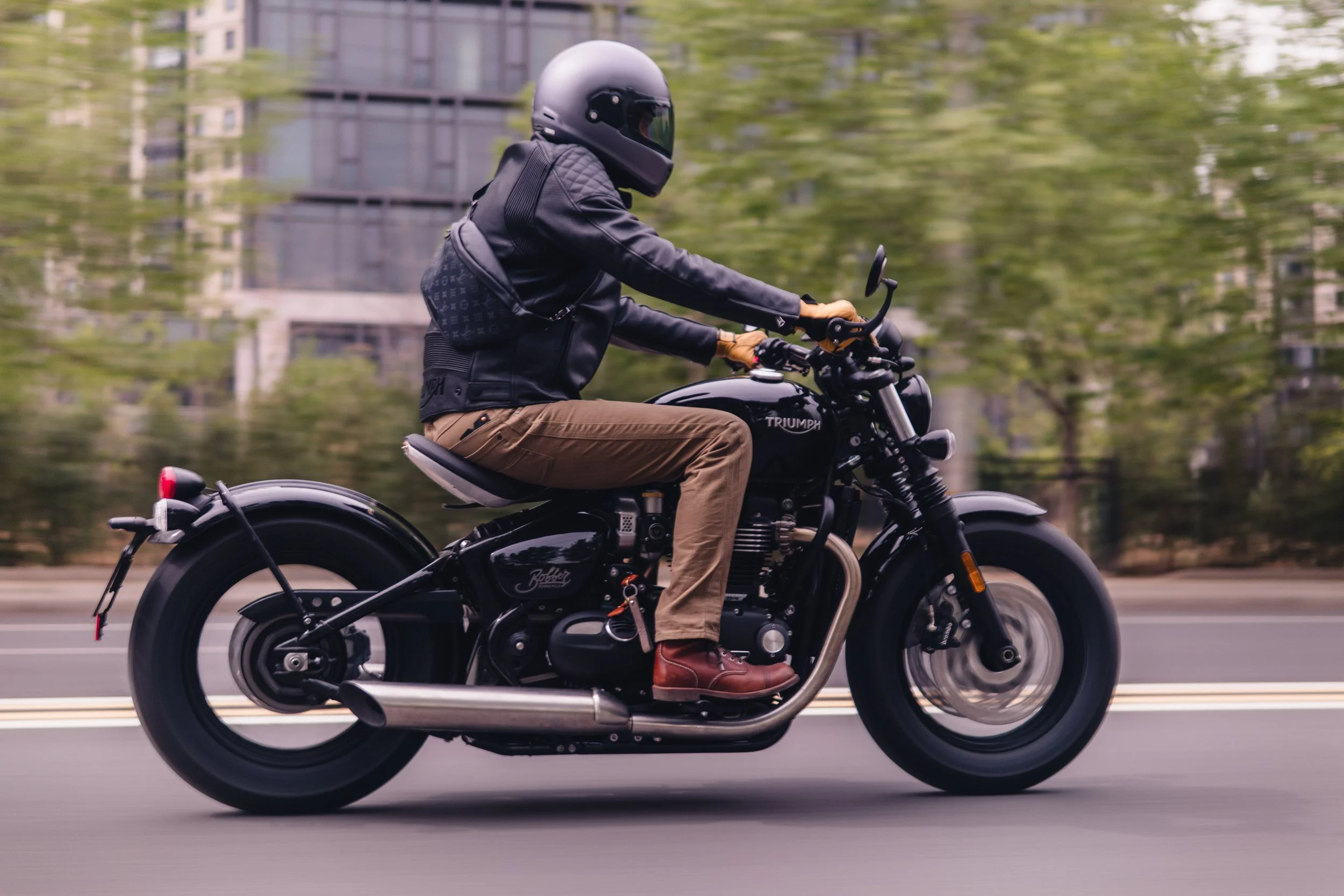 A person riding a black Triumph motorcycle on a city street, dressed in a black helmet, black leather jacket, tan pants, and brown boots, with a background of trees and buildings.