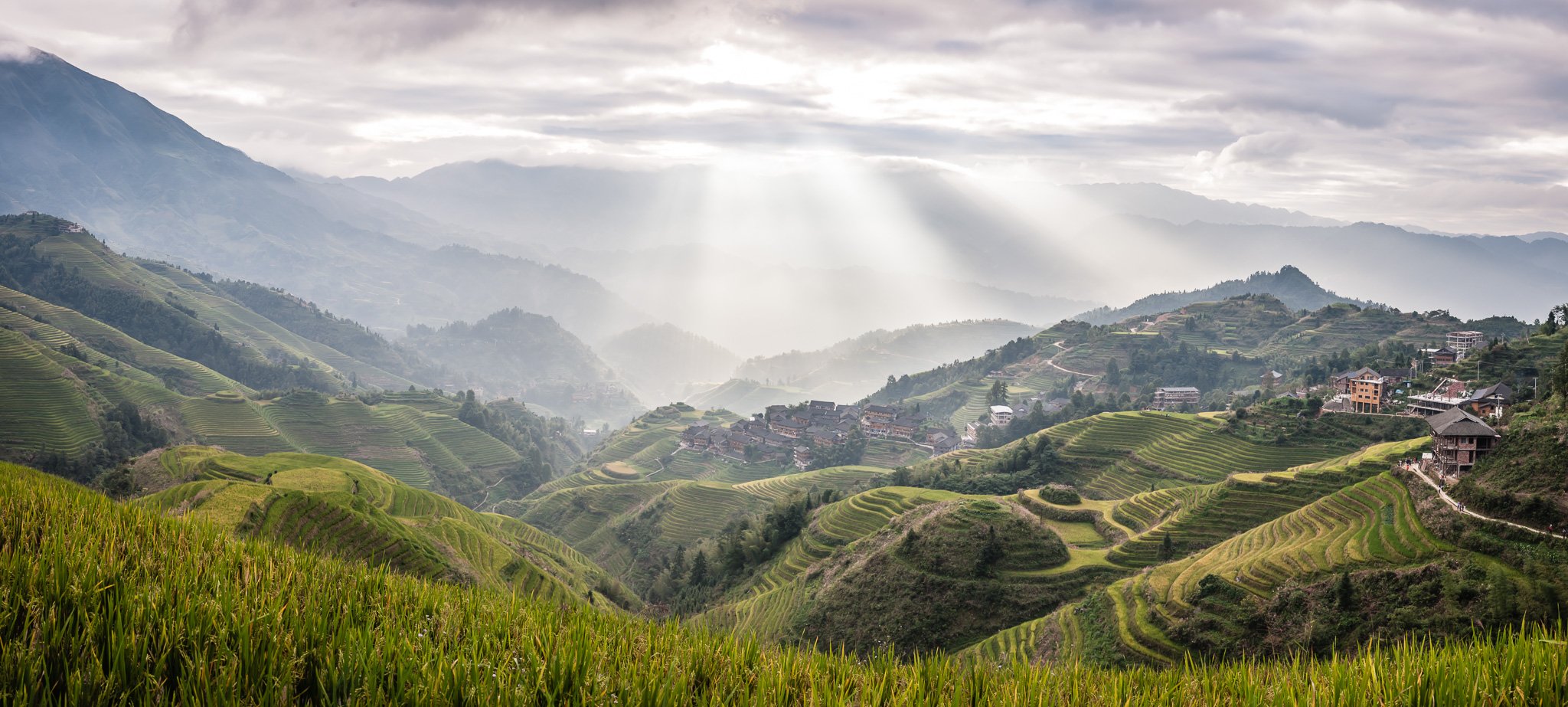 Terraced rice fields on rolling hills in a mountainous landscape with clouds and rays of sunlight breaking through, and a small village in the distance.