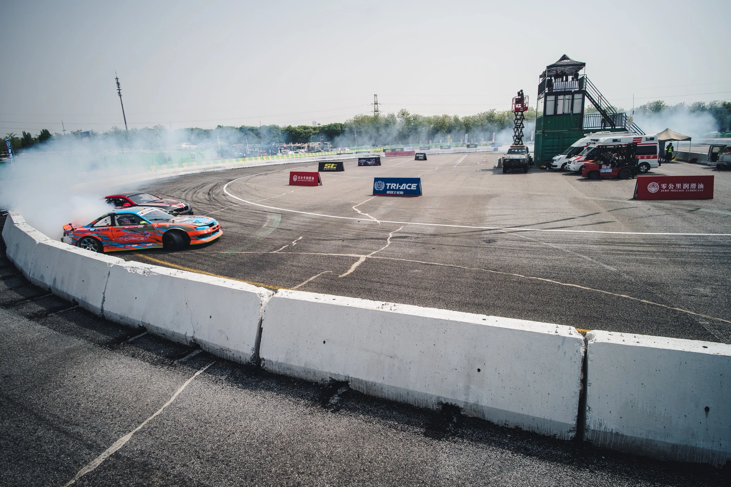 Race cars drifting around a corner on a racetrack with tire smoke, with barriers and track signage visible.