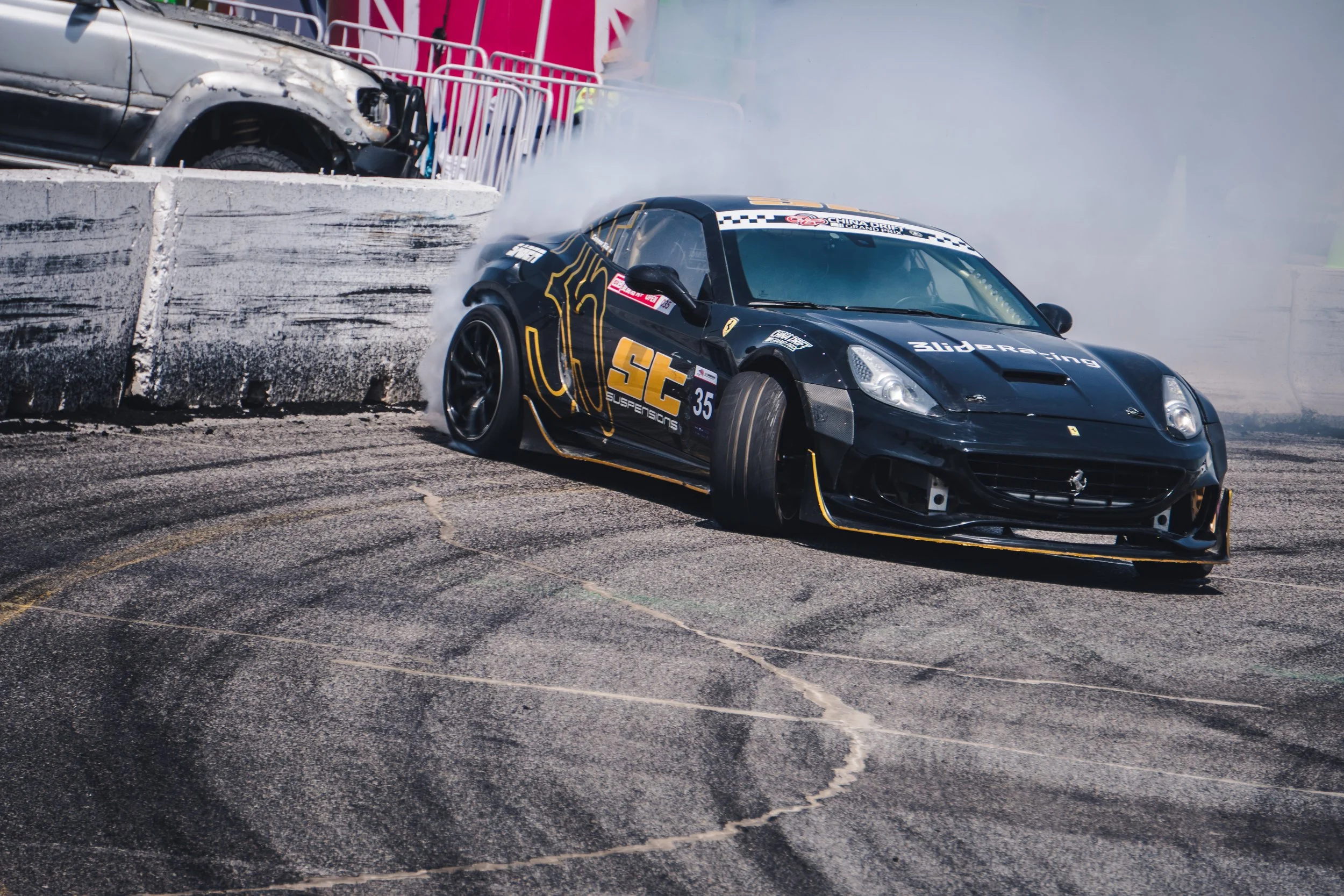 A black race car with yellow accents and the logo 'st suspensions' crashes into a concrete barrier during a motorsport event, creating smoke and tire skid marks on the asphalt.