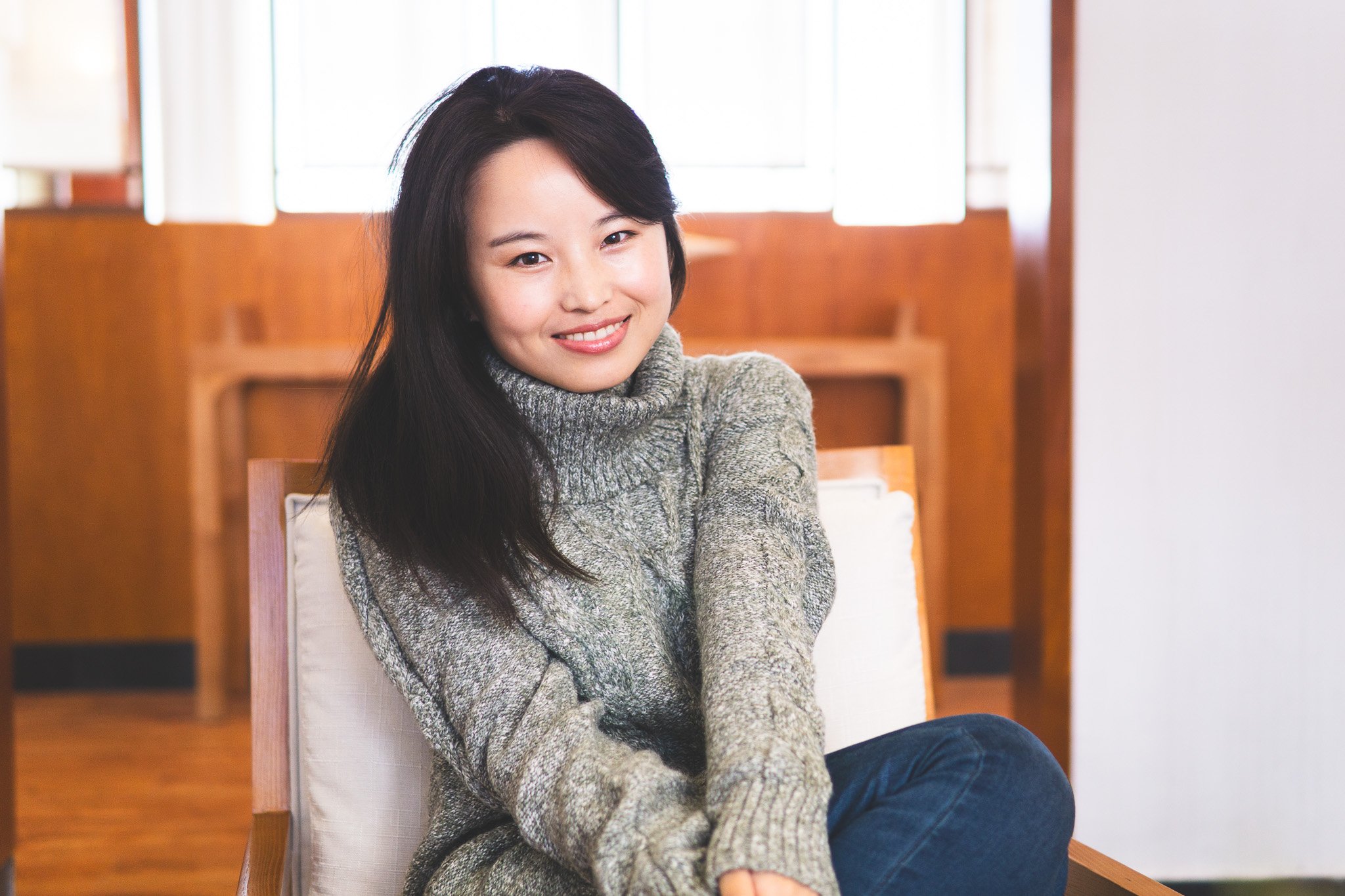A young woman with long black hair, smiling, wearing a gray turtleneck sweater, sitting in a wooden chair in a brightly lit room.