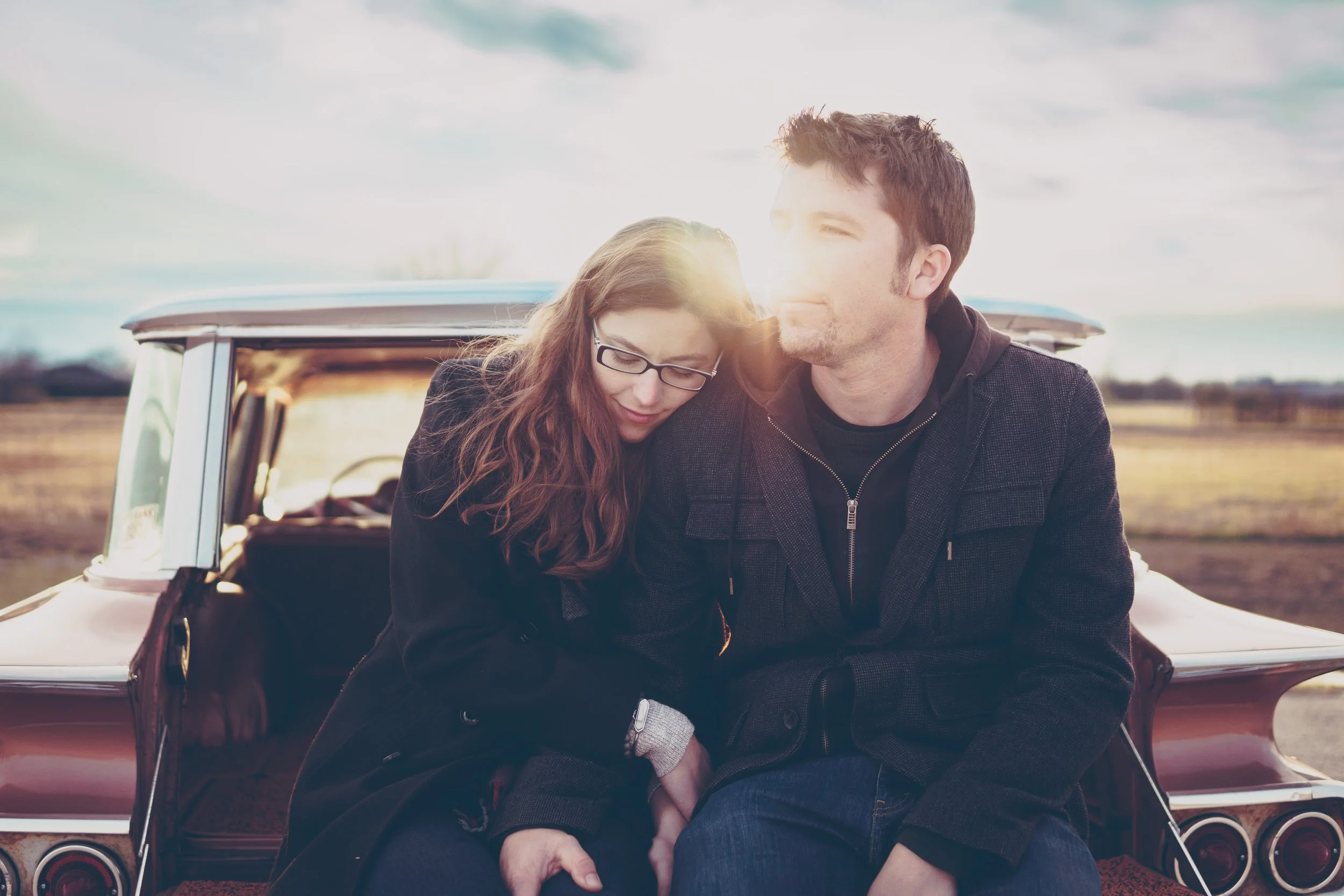 A man and woman sitting on the trunk of a vintage car in a field, with sunlight shining through, looking peaceful and close to each other.