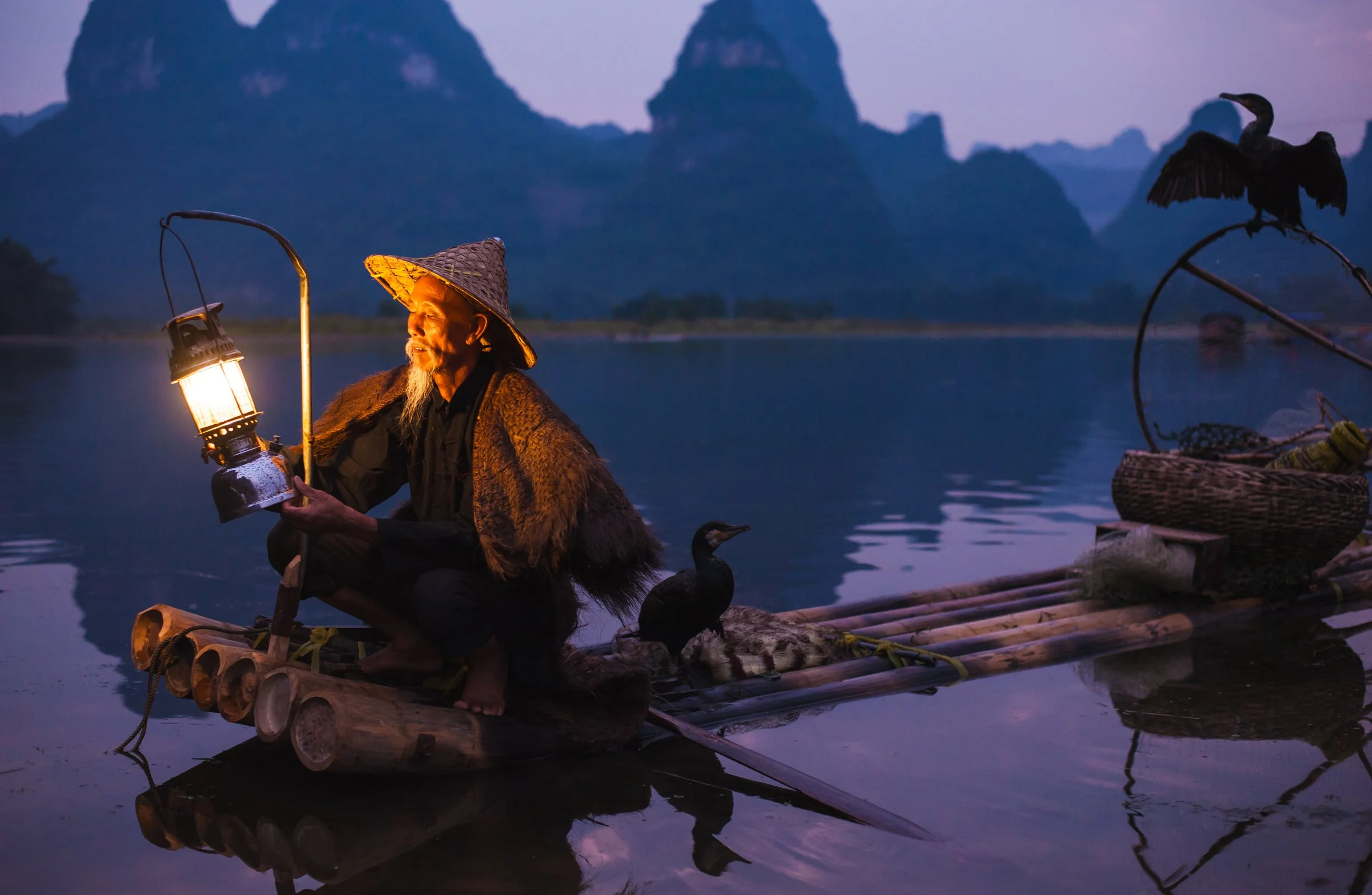 An elderly man with a traditional conical hat and fur cloak sitting on a bamboo raft on a lake, holding a lantern, with mountains in the background during dawn or dusk.