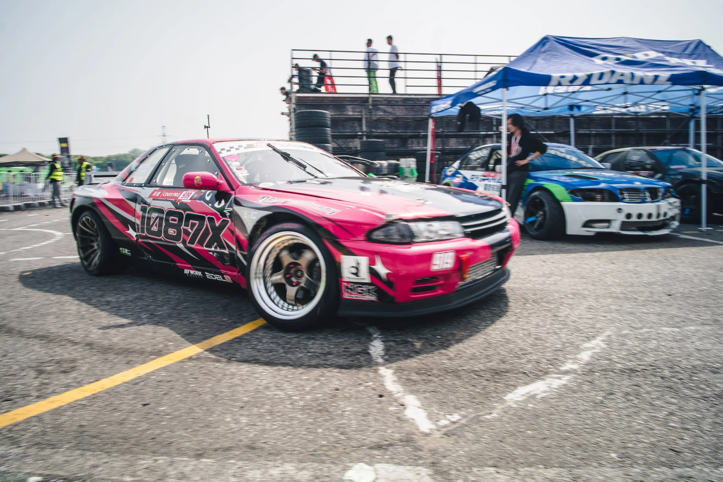 Pink and black race car in pit area with other cars and people in background at a racing event.