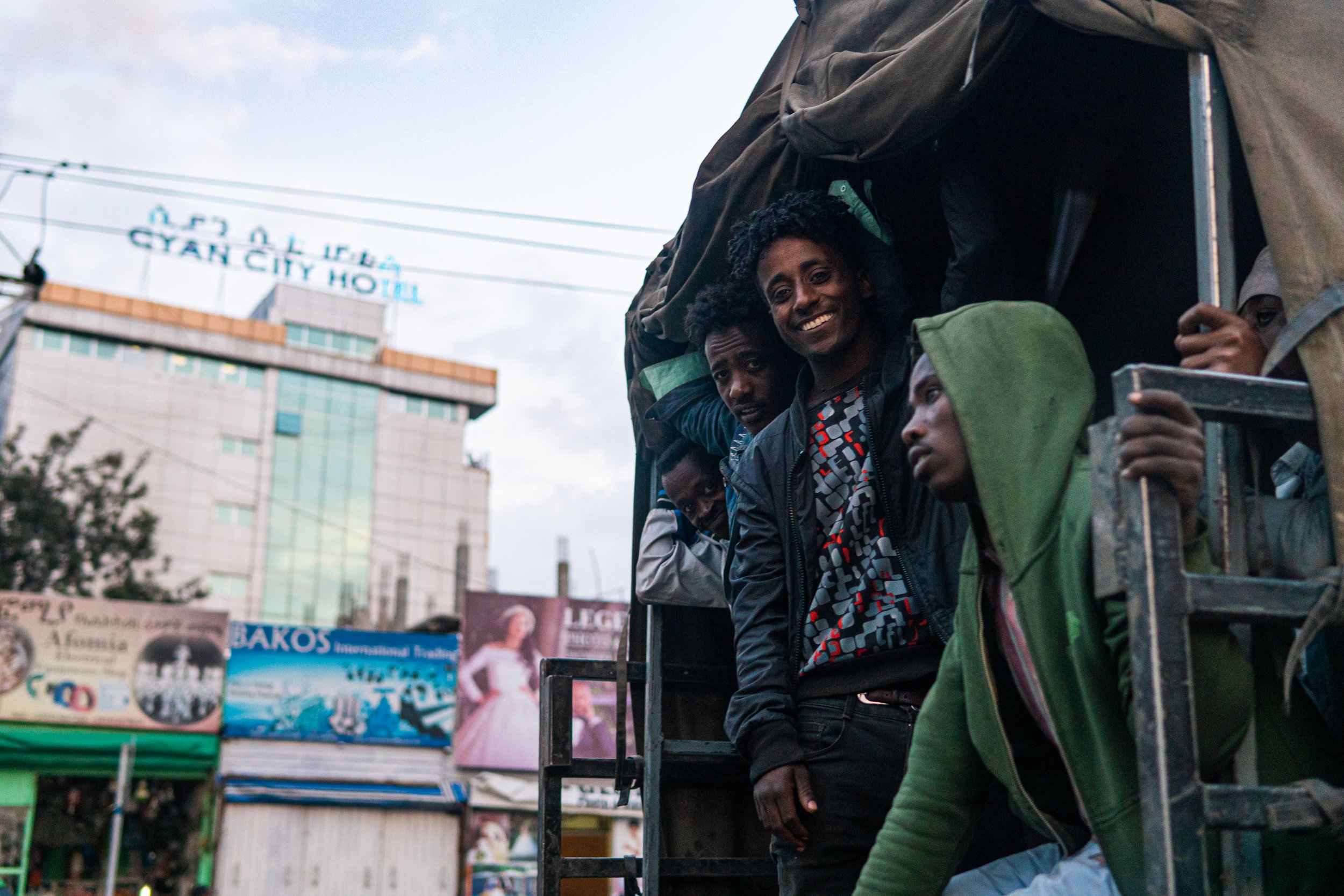 A group of men riding in the back of a truck on a city street with billboards and a hotel building in the background