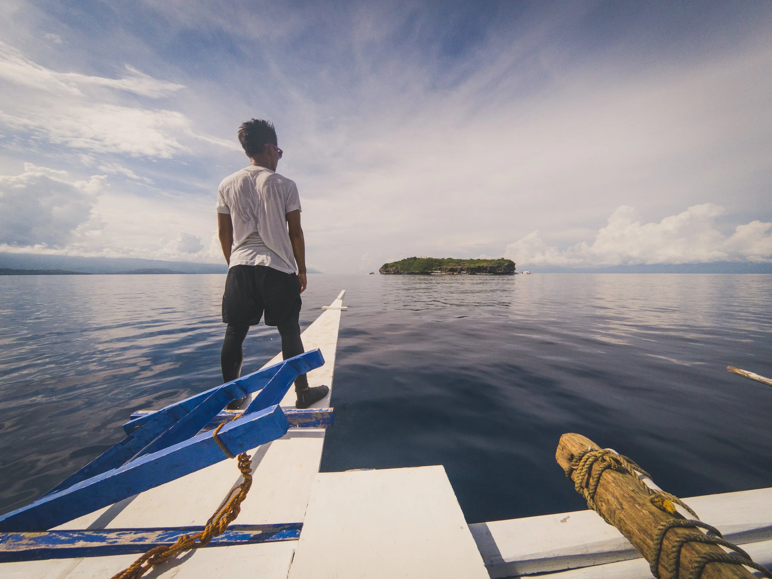 A person stands on the edge of a boat looking at an island in the distance on a calm body of water under a cloudy sky.