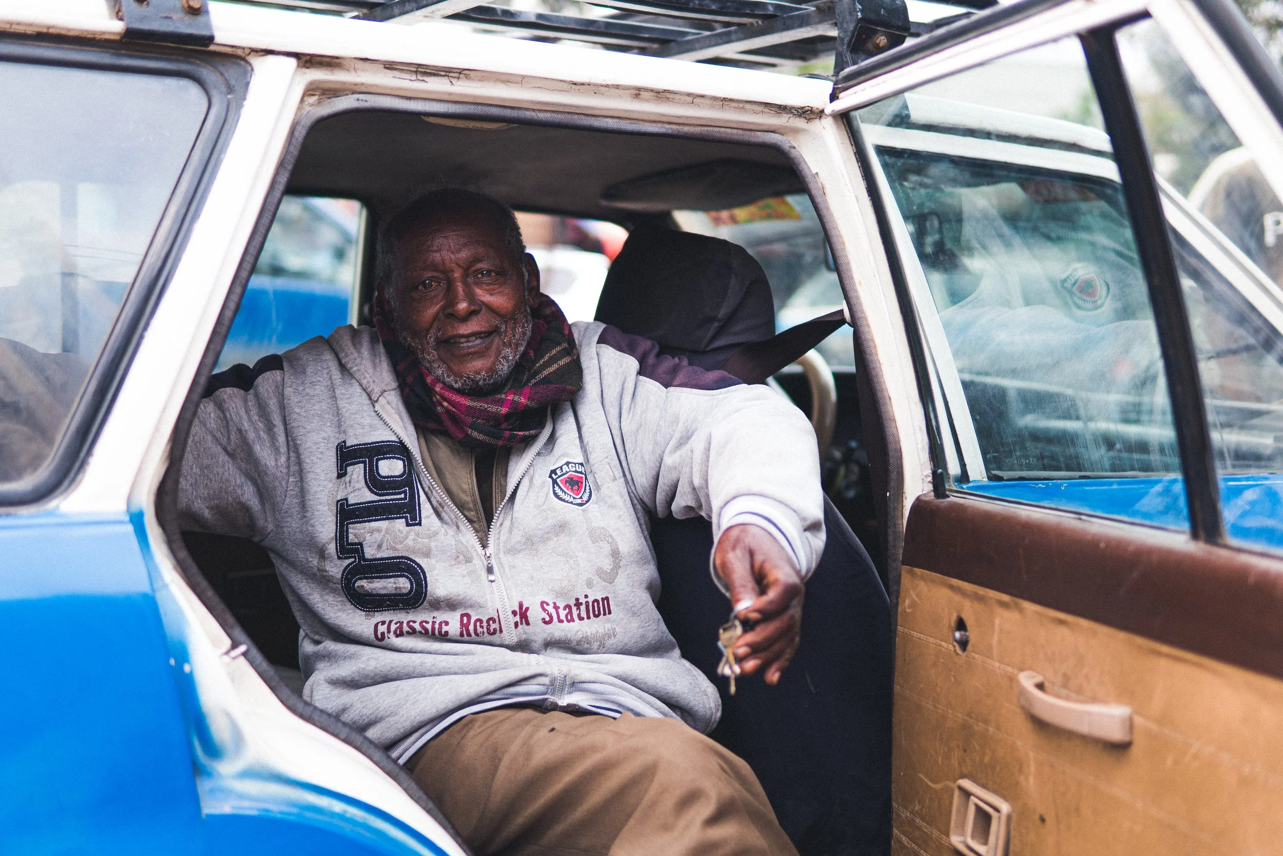 An elderly man sitting inside a blue and white vehicle, smiling, wearing a gray zip-up hoodie with police patch and 'Classic Rock Station' text, holding keys.