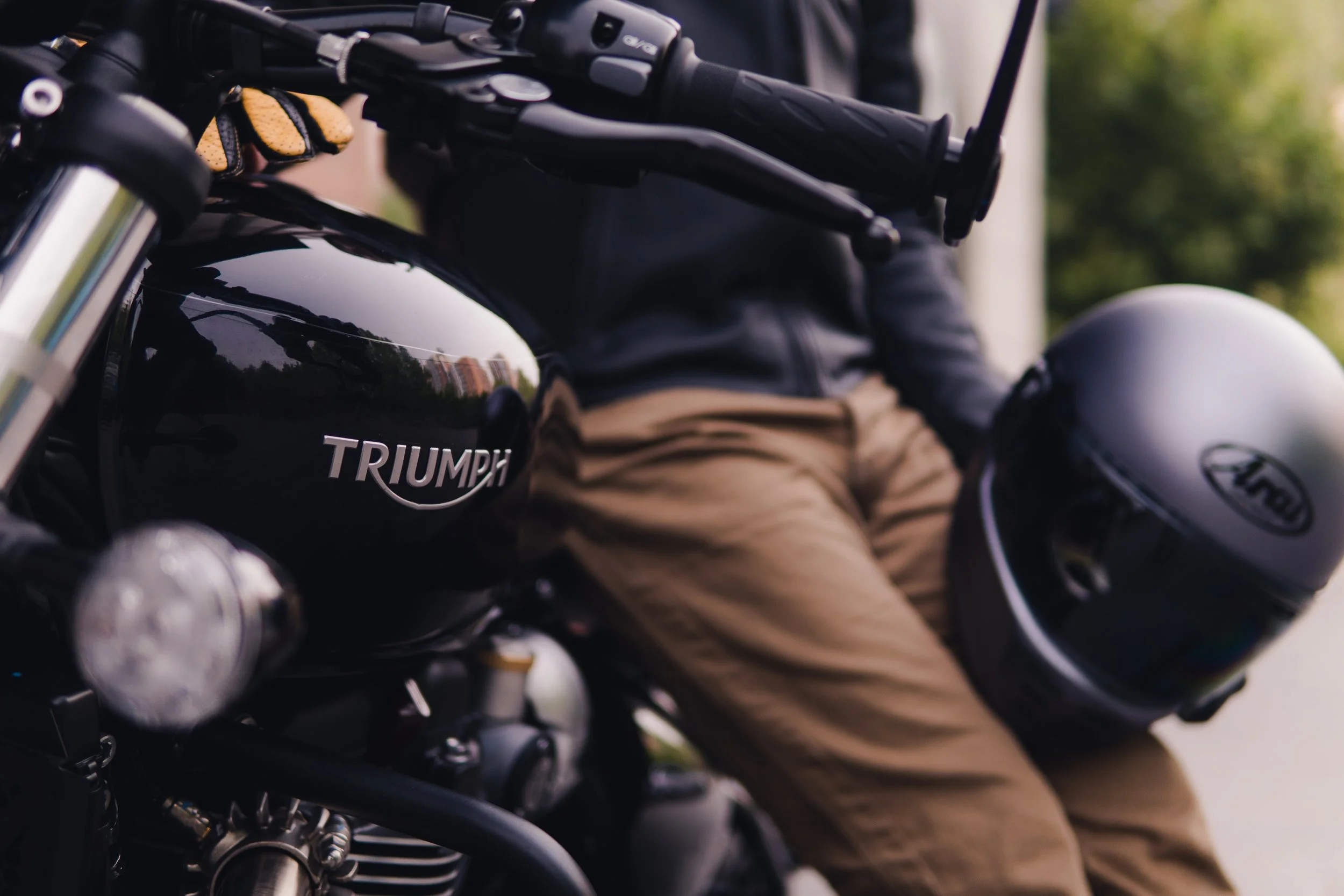 Close-up of a Triumph motorcycle with a person in leather jacket and tan pants sitting next to it, holding a black helmet with a logo, and the motorcycle's fuel tank and engine parts visible.