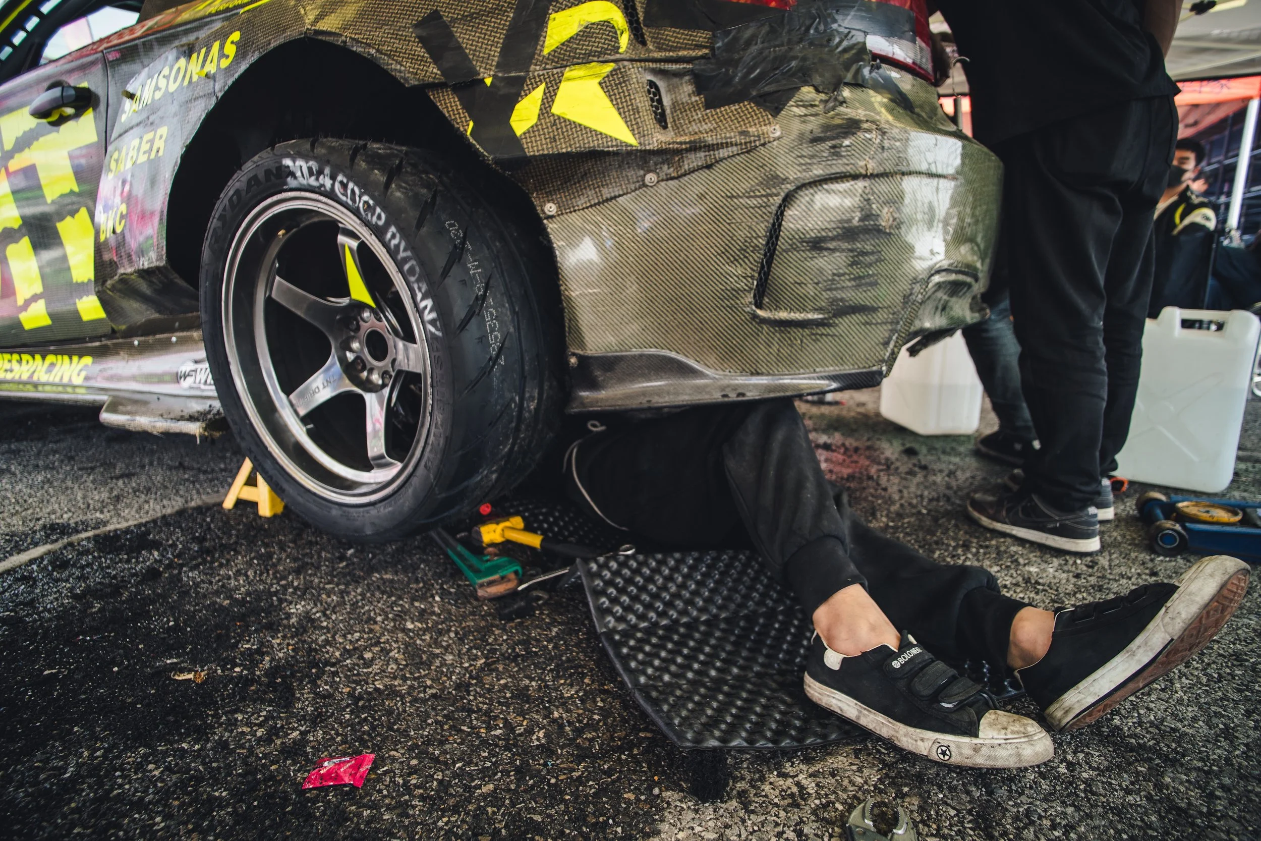Race car in a garage or pit area being serviced by a mechanic under the vehicle, with various tools and equipment around.