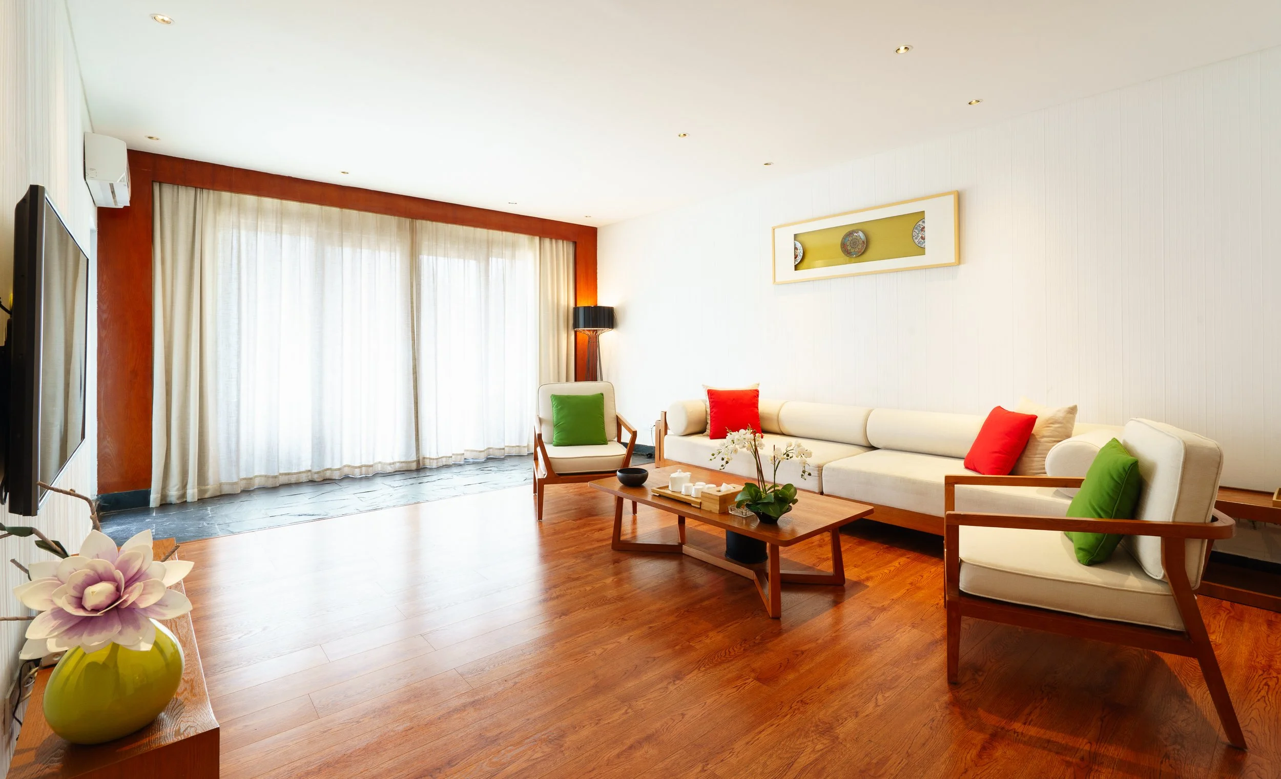 Living room with white sofa, armchairs with green and red pillows, a wooden coffee table with orchids, and large windows with cream curtains.