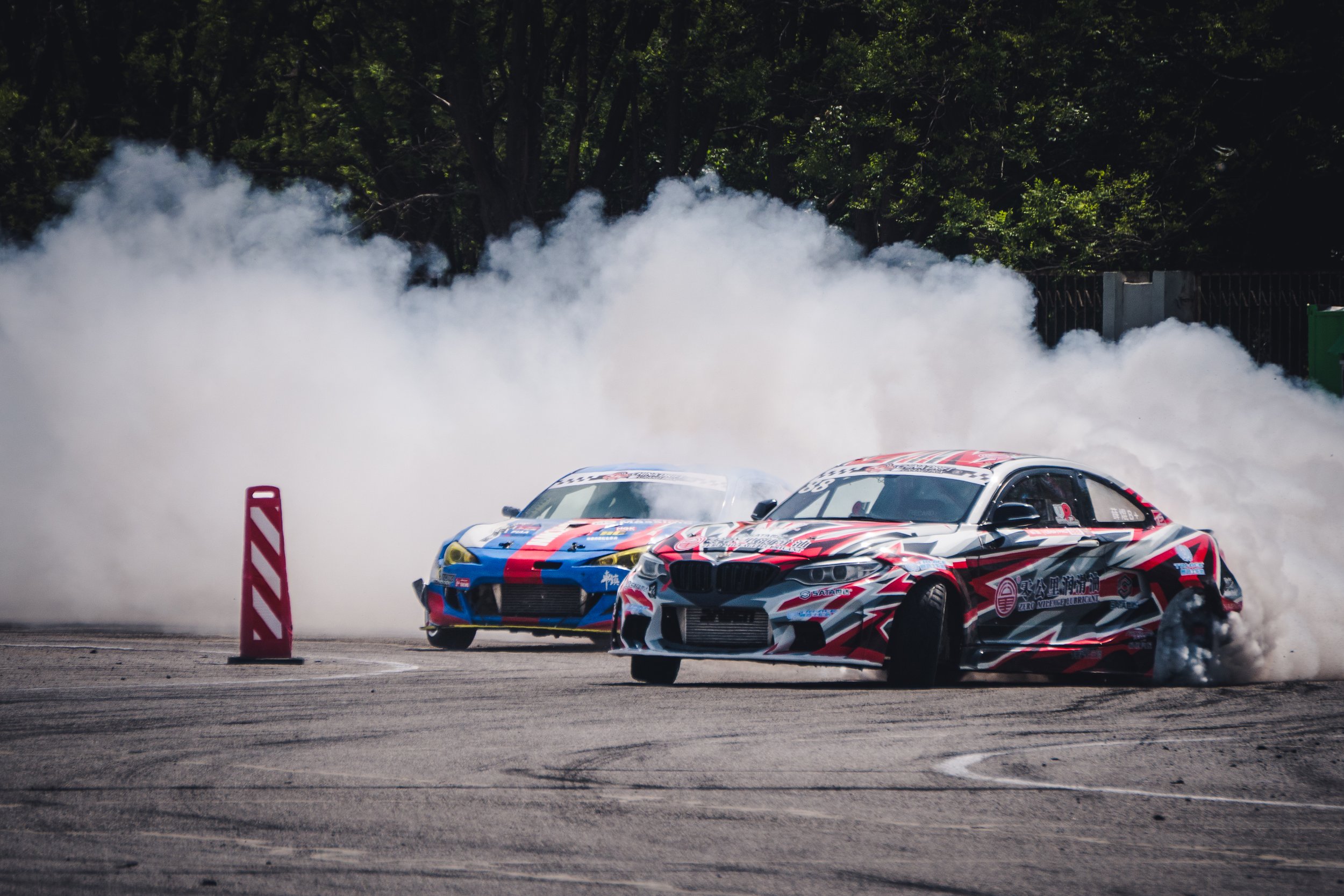Two race cars drifting on a track with smoke billowing behind them, a red and white striped barrier to the left, and green trees in the background.