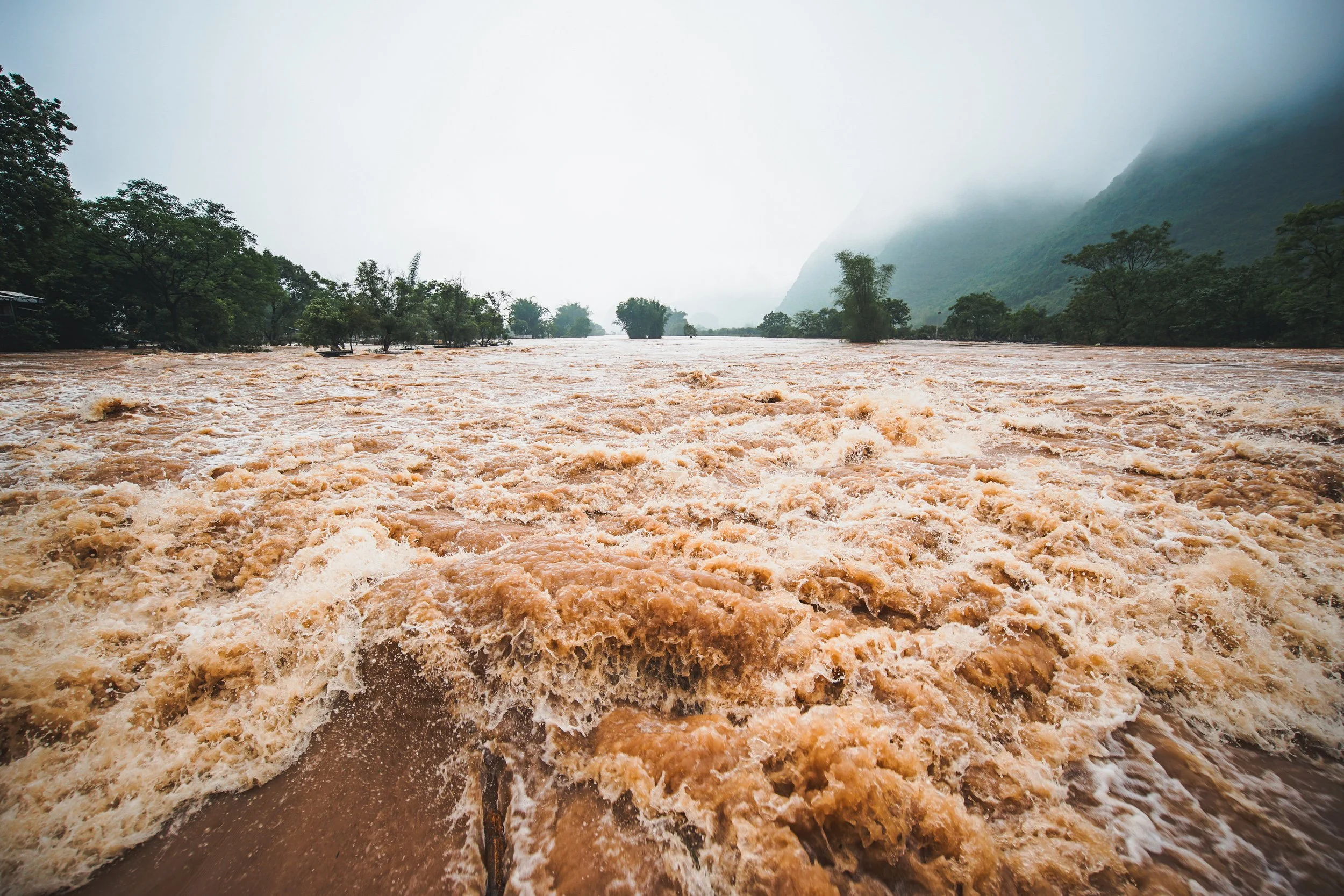 Flooded river with muddy water, surrounded by trees and mountains, under cloudy and misty sky.