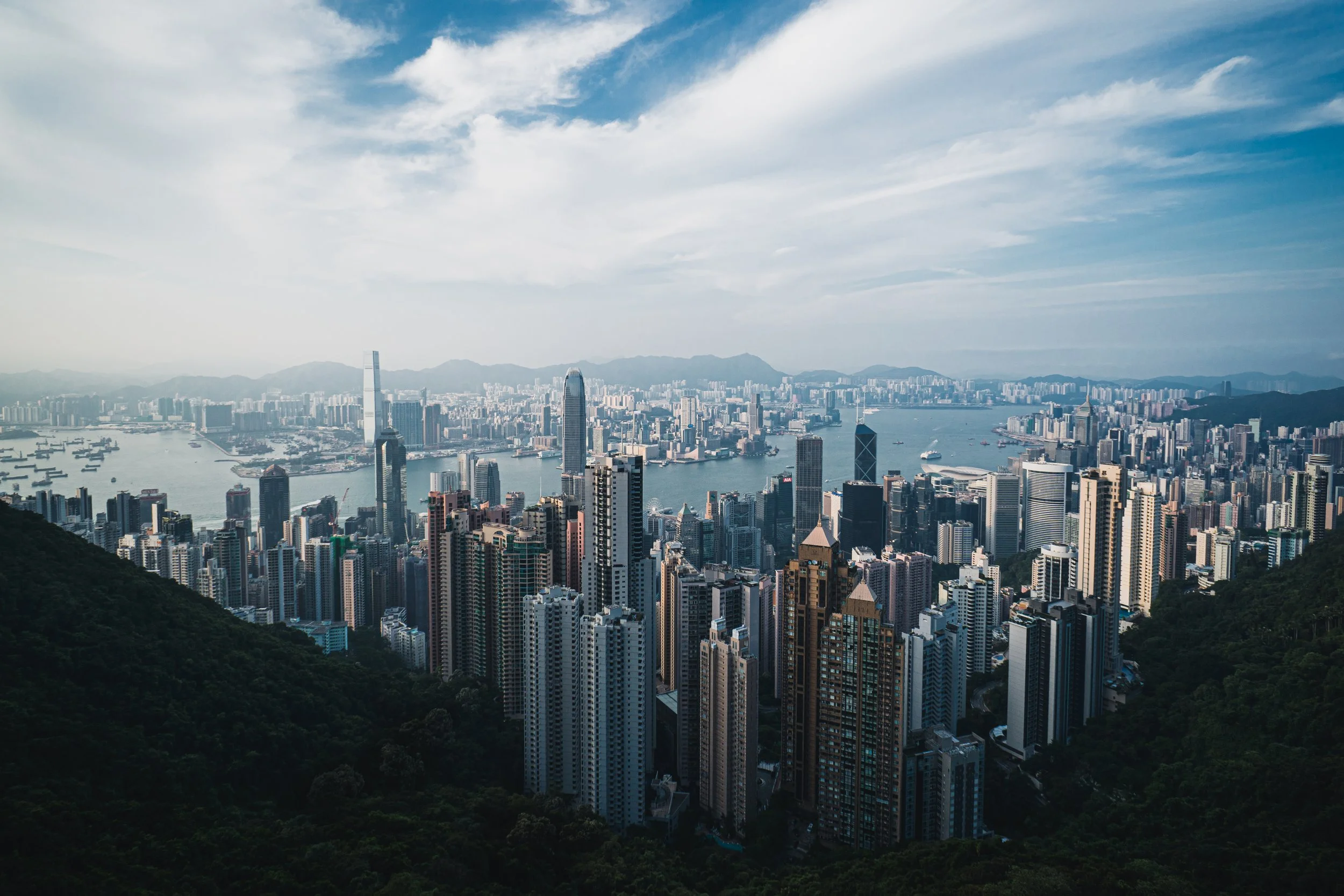 A city skyline with numerous high-rise buildings, a body of water with boats, and mountains in the background under a partly cloudy sky.