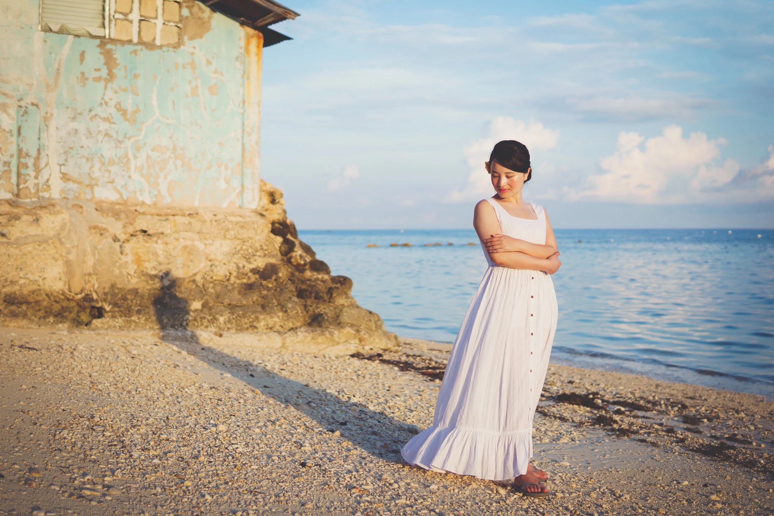A woman in a long white dress standing on a sandy beach near the water with an old weathered building in the background, under a partly cloudy sky.