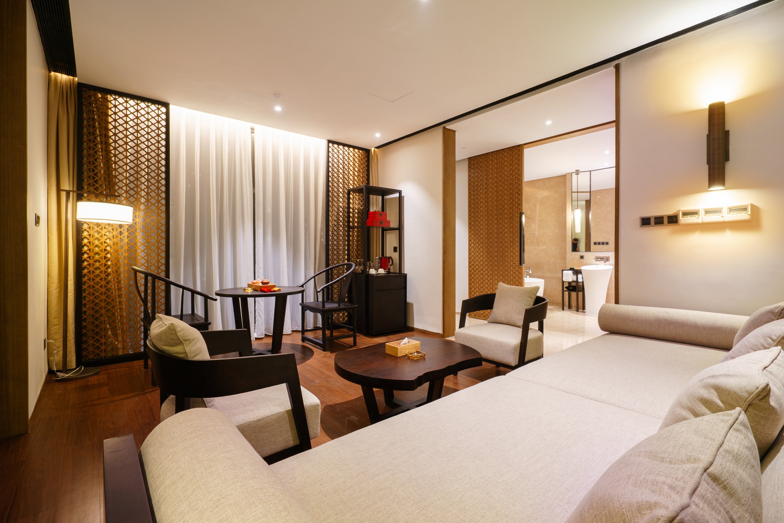 Living room with beige sofa, armchair, small round table, and a black coffee table. In the background, there is a small table with a tea set and a shelf with red items. The room has wooden flooring, beige walls, and sheer white curtains covering wind
