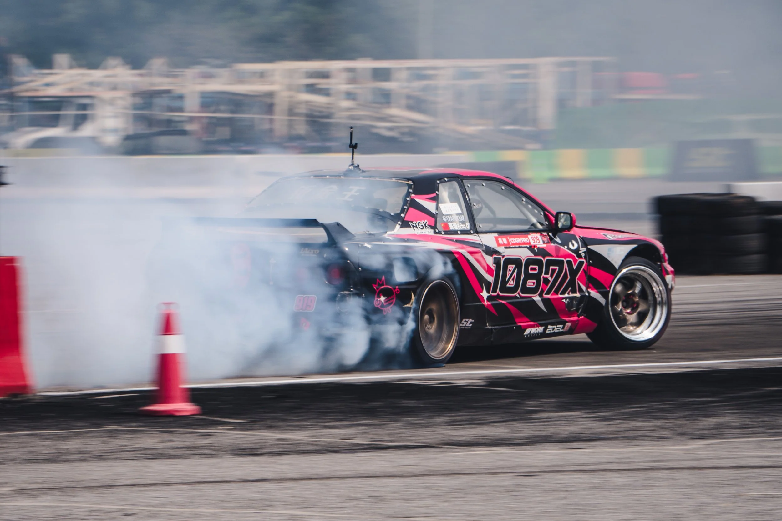 A black and pink race car drifting on a track with tire smoke, orange cones, and blurred background.