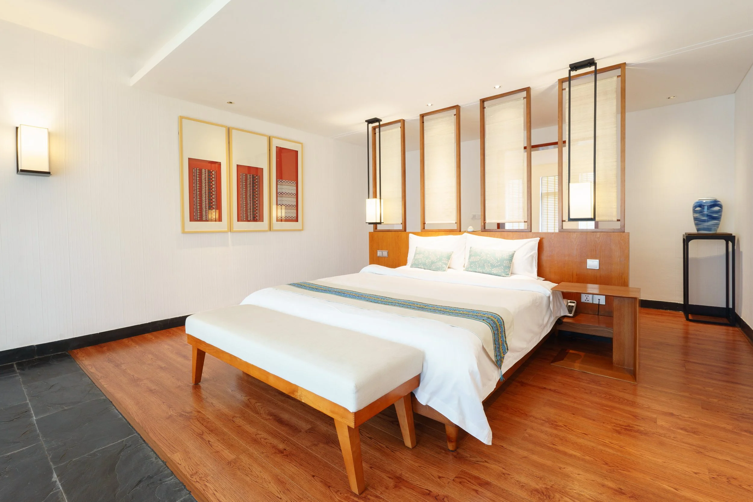 A modern hotel room with a large bed, a wooden headboard, a bench at the foot of the bed, and decorative artwork on the wall. There are partition panels behind the bed, hanging lights, and a small table with a blue vase.