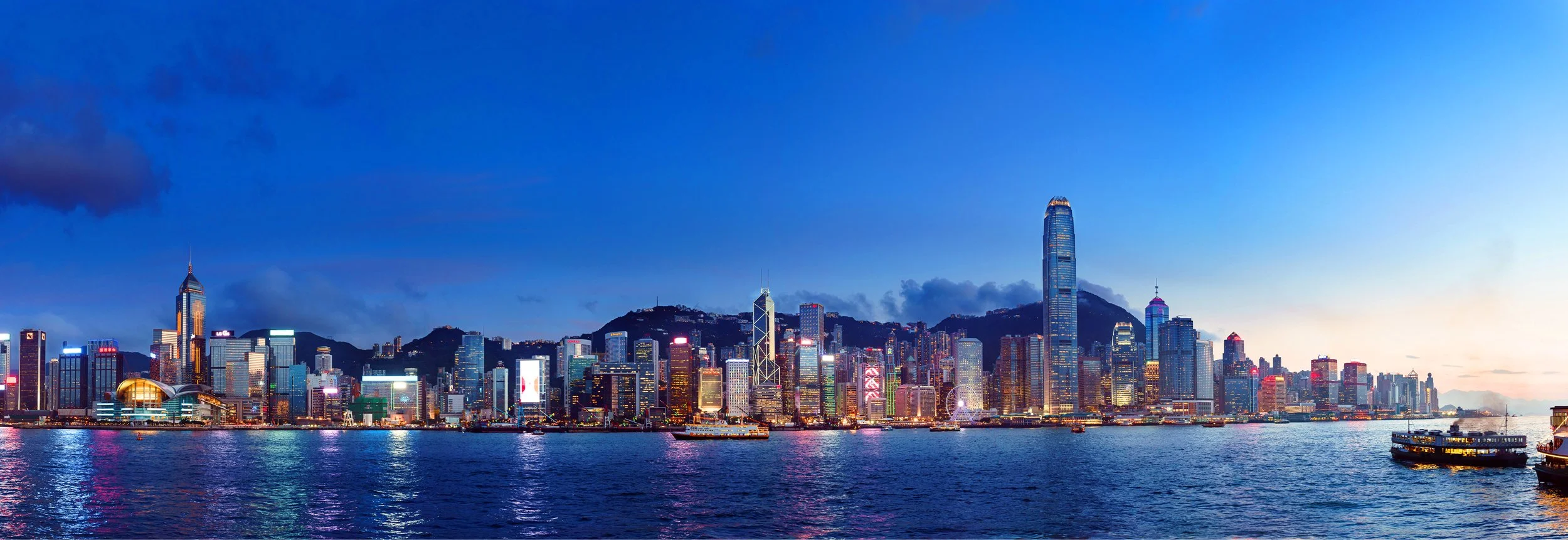Nighttime view of a city skyline across a body of water, with illuminated skyscrapers and mountains in the background.