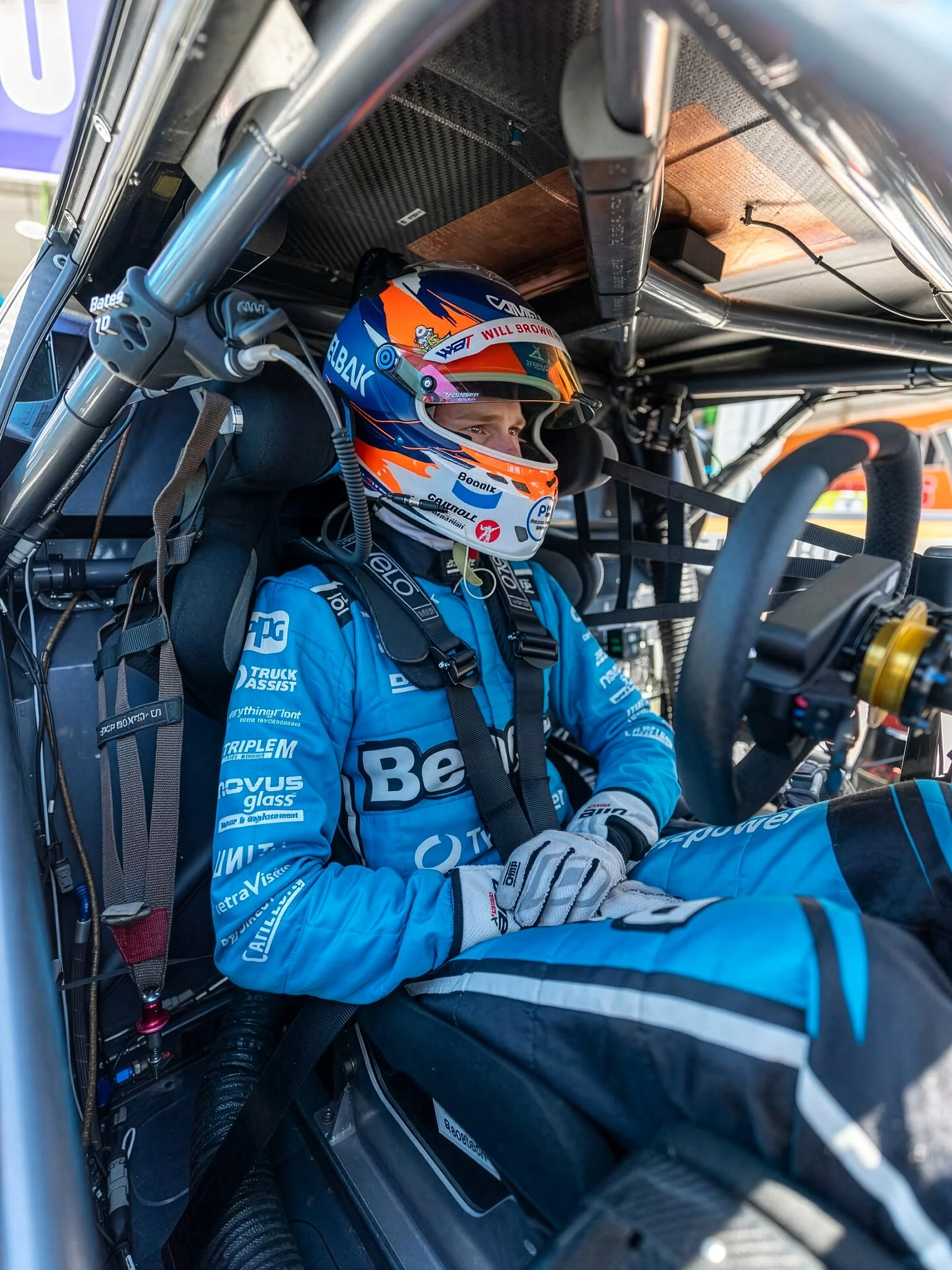 A race car driver in a blue racing suit and helmet sitting inside a race car cockpit, preparing for a race or practice session.