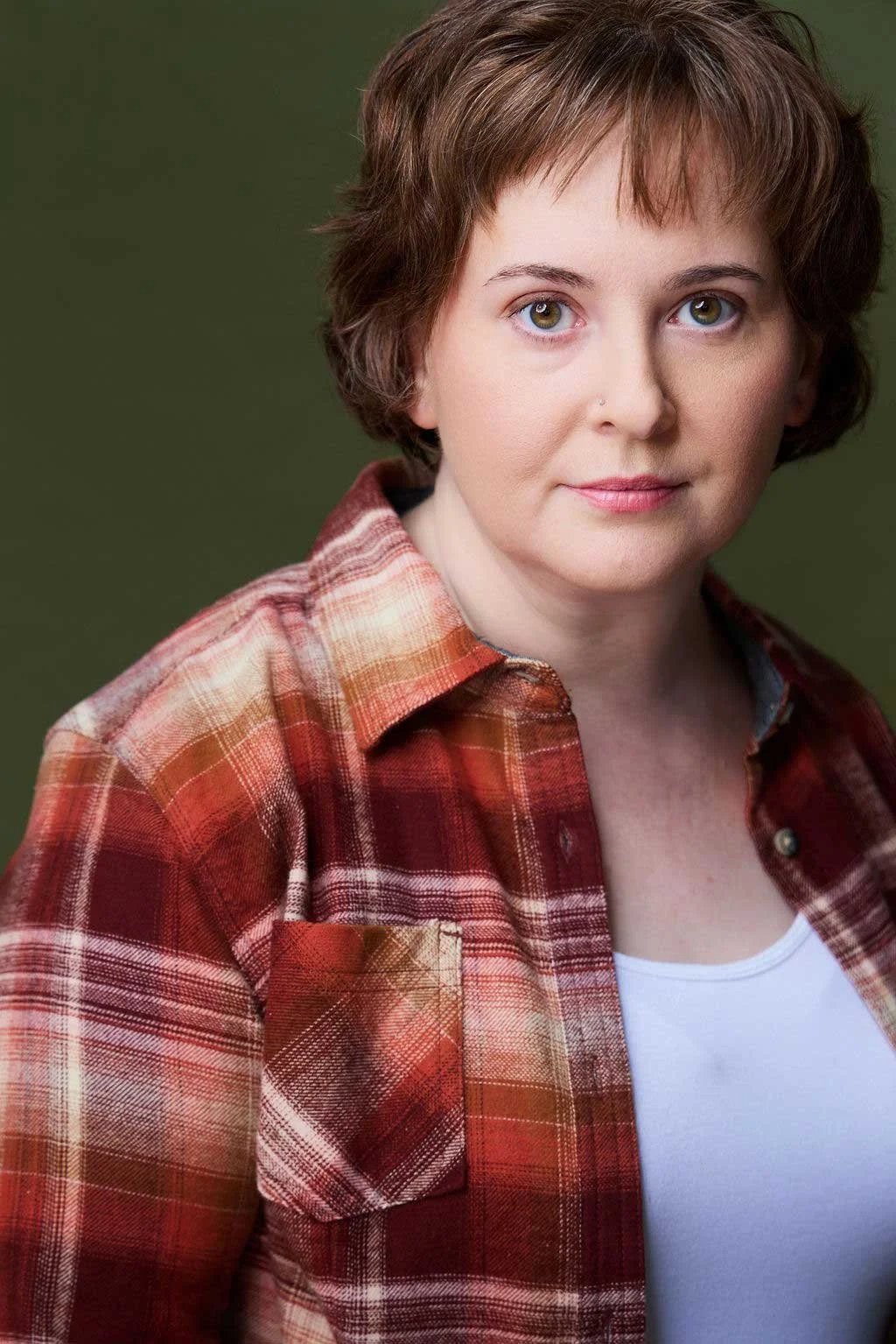 Sue Gauthier has her brown hair down, a closed-mouth smile, looking at the camera. She's wearing a red-and-orange plaid button-up shirt, showing her range as an actress/writer.