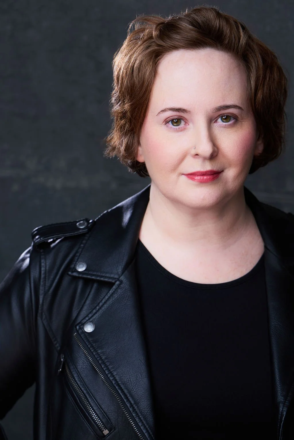 Sue Gauthier has her brown hair down, a closed-mouthed smirk, looking at the camera. Wearing a black leather jacket and a black undershirt, showcasing her range as an actress/writer.