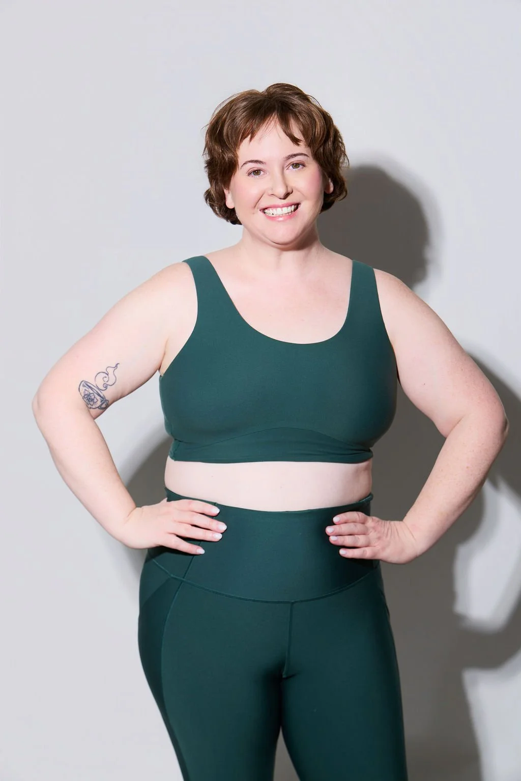 Sue Gauthier, with brown hair, wears a forest-green workout set, smiling at the camera, her hands on her hips. She showcases her roles as an actress/writer.