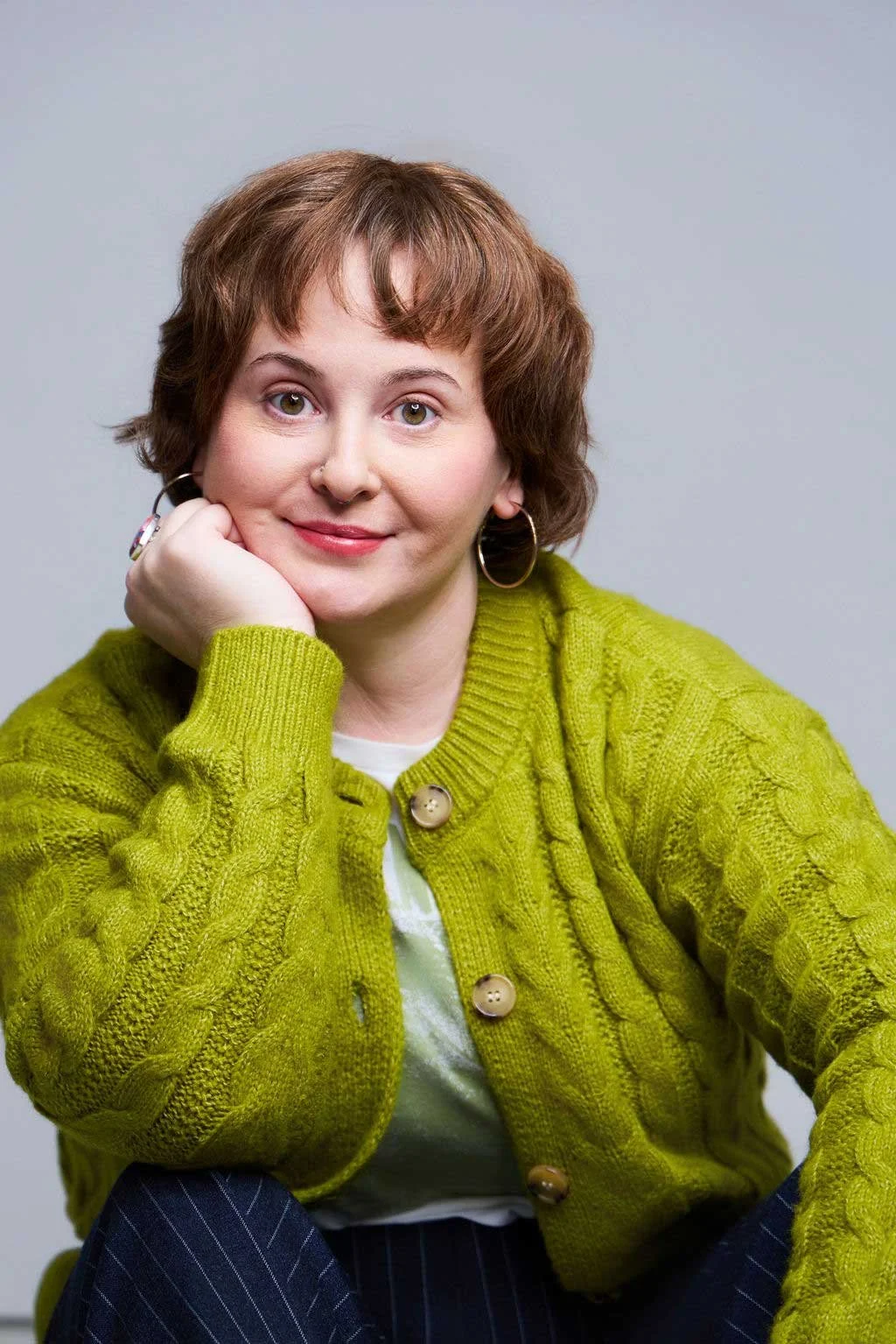 Sue Gauthier has brown hair down, smiles with closed lips, and wears gold hoop earrings. She’s in a green sweater, cardigan, and white undershirt, showcasing her versatility as an actress and writer.