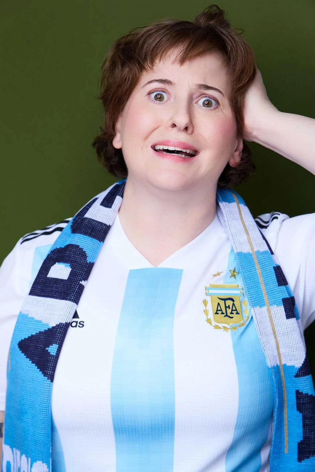 Sue Gauthier, with brown hair, wears a sports jersey and a scarf, expressing a stressed facial expression, looking at the camera. She showcases her roles as an actress/writer.