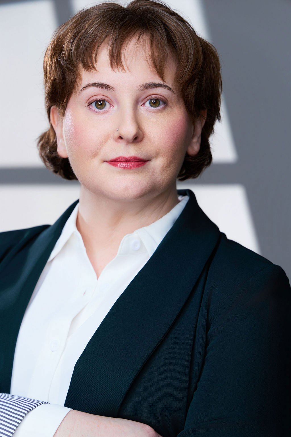 Sue Gauthier has her brown hair down, a serious smirk, and her arms crossed. Wearing a black business jacket and white button-down shirt, showing her range as an actress/writer.