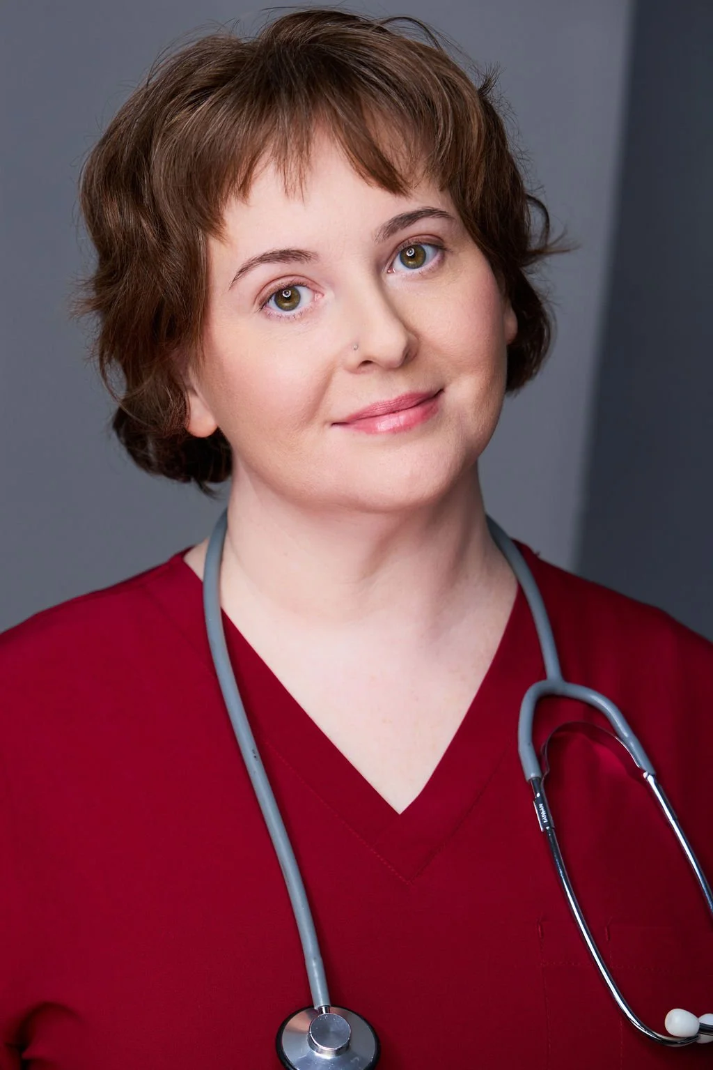 Sue Gauthier has her brown hair down, a welcoming smile, and a stethoscope around her neck. Wearing red scrubs, showing her range as an actress/writer.