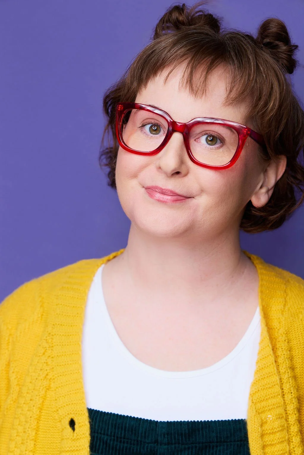 Sue Gauthier, with brown hair in mini buns and red glasses, wears a bright yellow cardigan and white undershirt, smiling at the camera. She showcases her roles as an actress/writer.