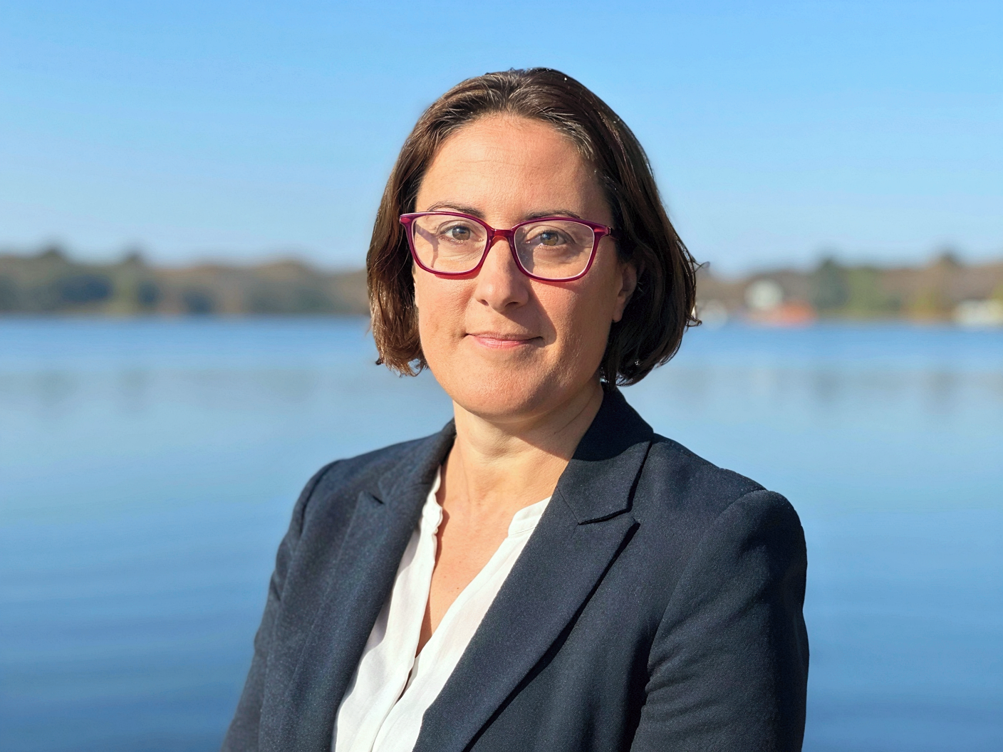 A woman with short brown hair and pink glasses standing outdoors by a body of water during daytime.