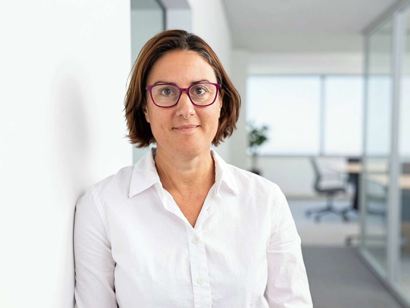A woman with short brown hair wearing glasses and a white shirt in an office setting with large windows