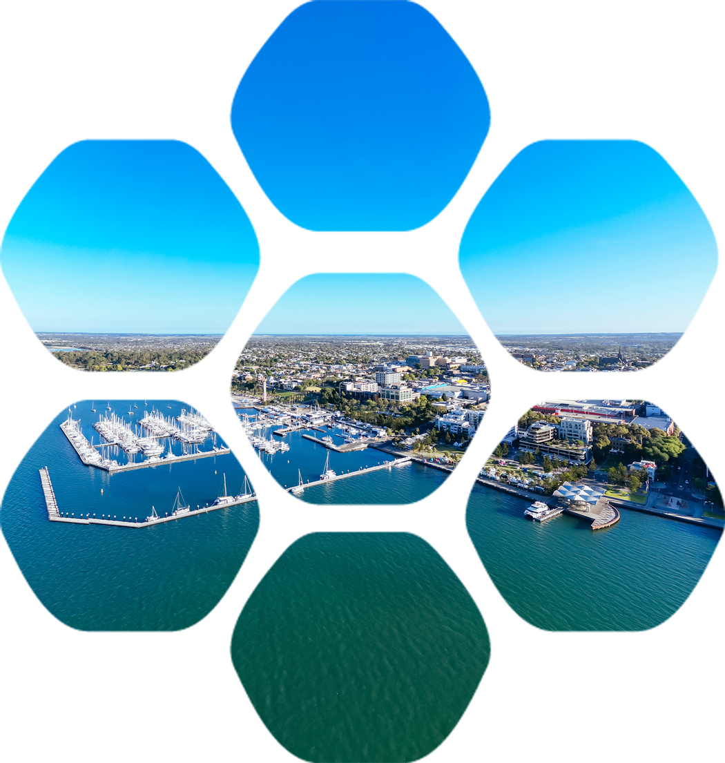 Aerial view of a marina with boats docked, surrounding buildings, and a cityscape in the distance under a clear blue sky.