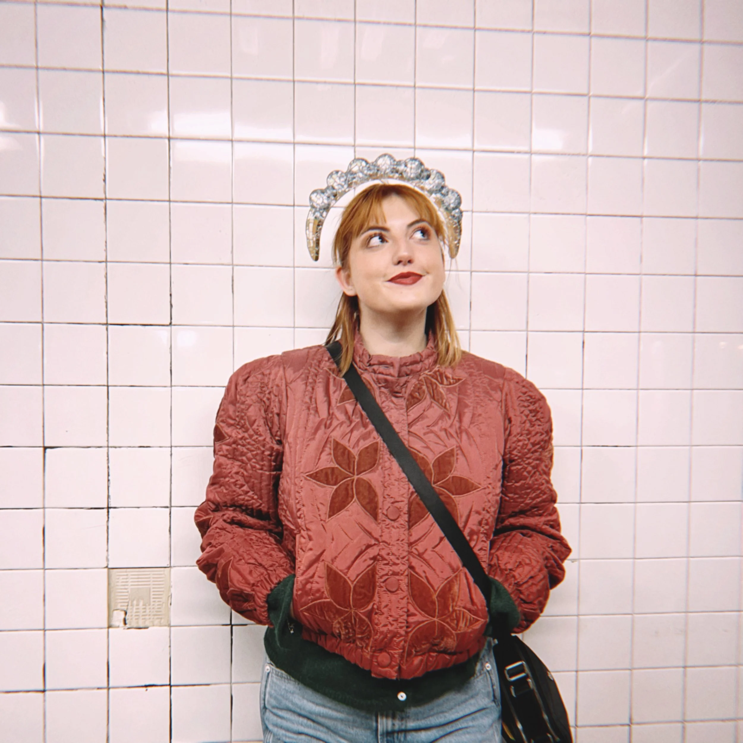 A young woman with red hair wearing a jeweled headband and a red embroidered jacket standing against a tiled wall.