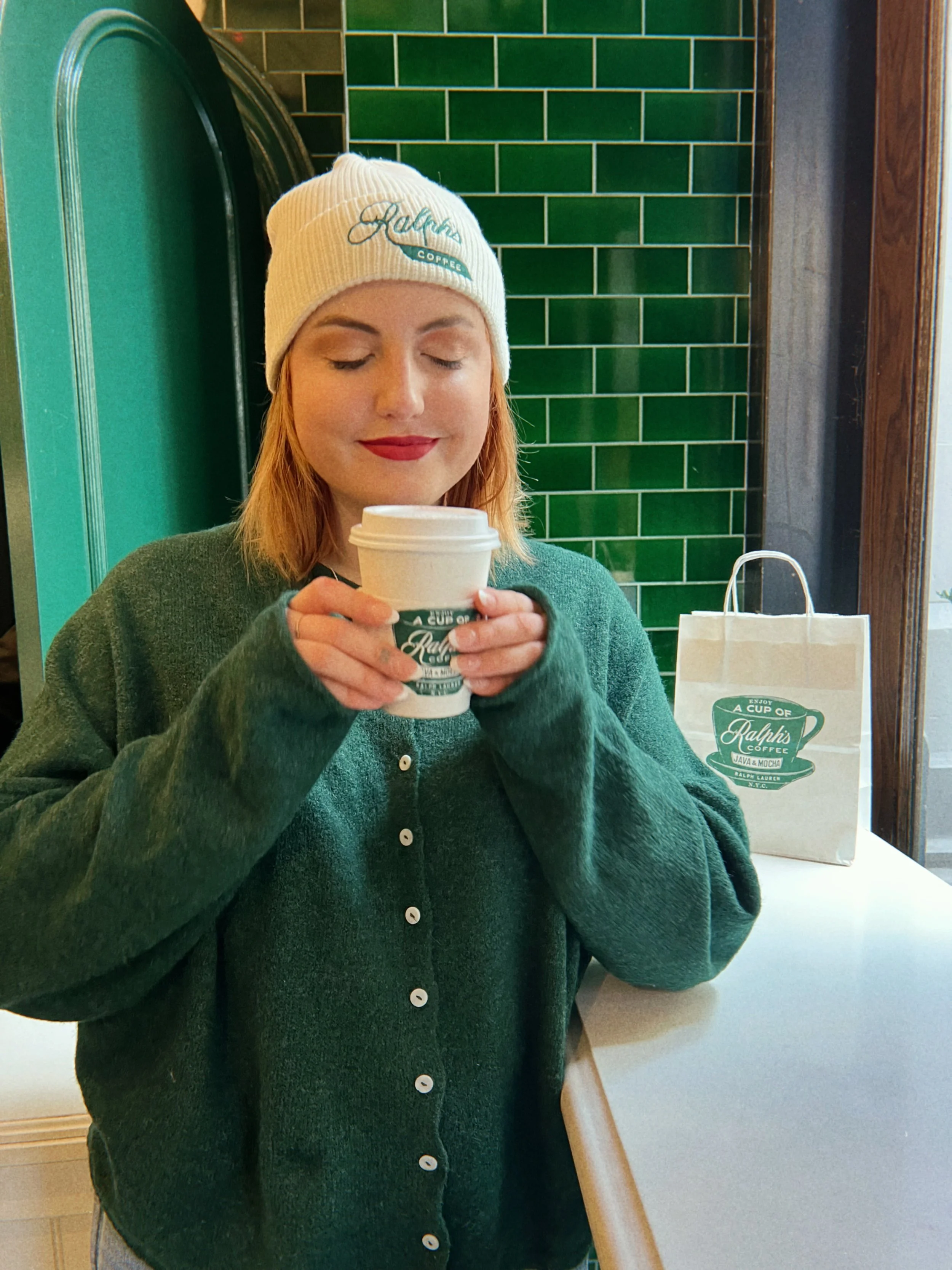 A young woman with red hair, wearing a white Ralphs beanie and a green sweater, holds a coffee cup in a coffee shop with green tiled walls and a paper Ralphs bag on the counter.