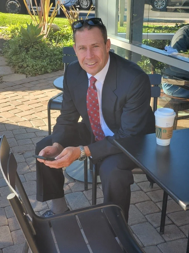 A man in a black suit, white shirt, and red patterned tie sitting outside at a table at Starbucks, holding a smartphone, with a cup of coffee on the table.
