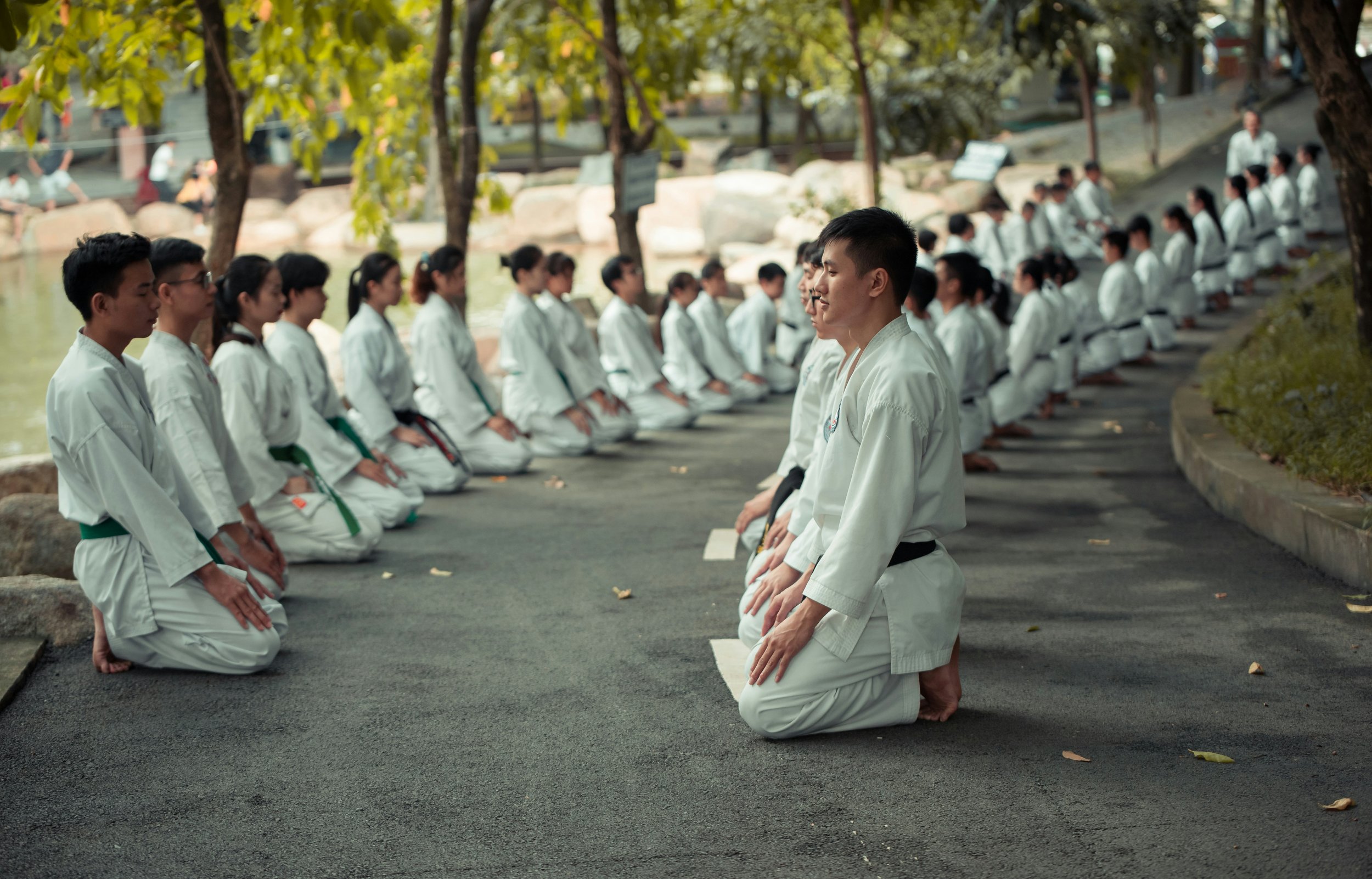 A group of martial artists dressed in white uniforms with black or green belts, kneeling on a paved path beside a lake, practicing meditation or prayer in an outdoor setting with trees and rocks in the background.
