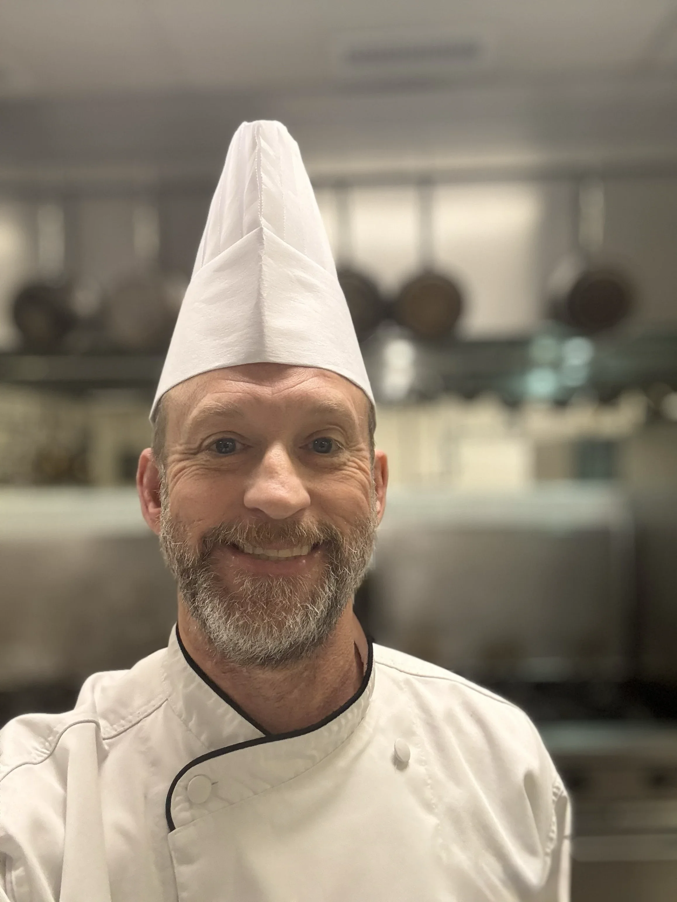 A smiling chef taking a selfie in a professional kitchen, wearing a white chef's coat and a tall white chef's hat.