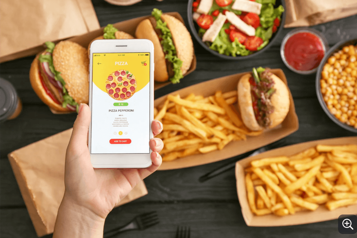 Hand holding a smartphone with online food ordering app open, displaying a pizza labeled 'Pizza Pepperoni' with a price of $12. Surrounding the phone are fast food items on a dark wooden table, including three sandwiches with lettuce and other fillings, two baskets of French fries, a bowl of salad with lettuce, cherry tomatoes, and cheese, a small bowl of ketchup, and a bowl of chickpeas.