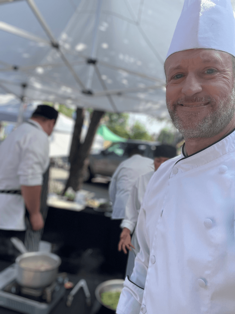 Smiling chef in a white chef's coat and hat taking a selfie outdoors, with other chefs and cooking stations visible under a canopy.