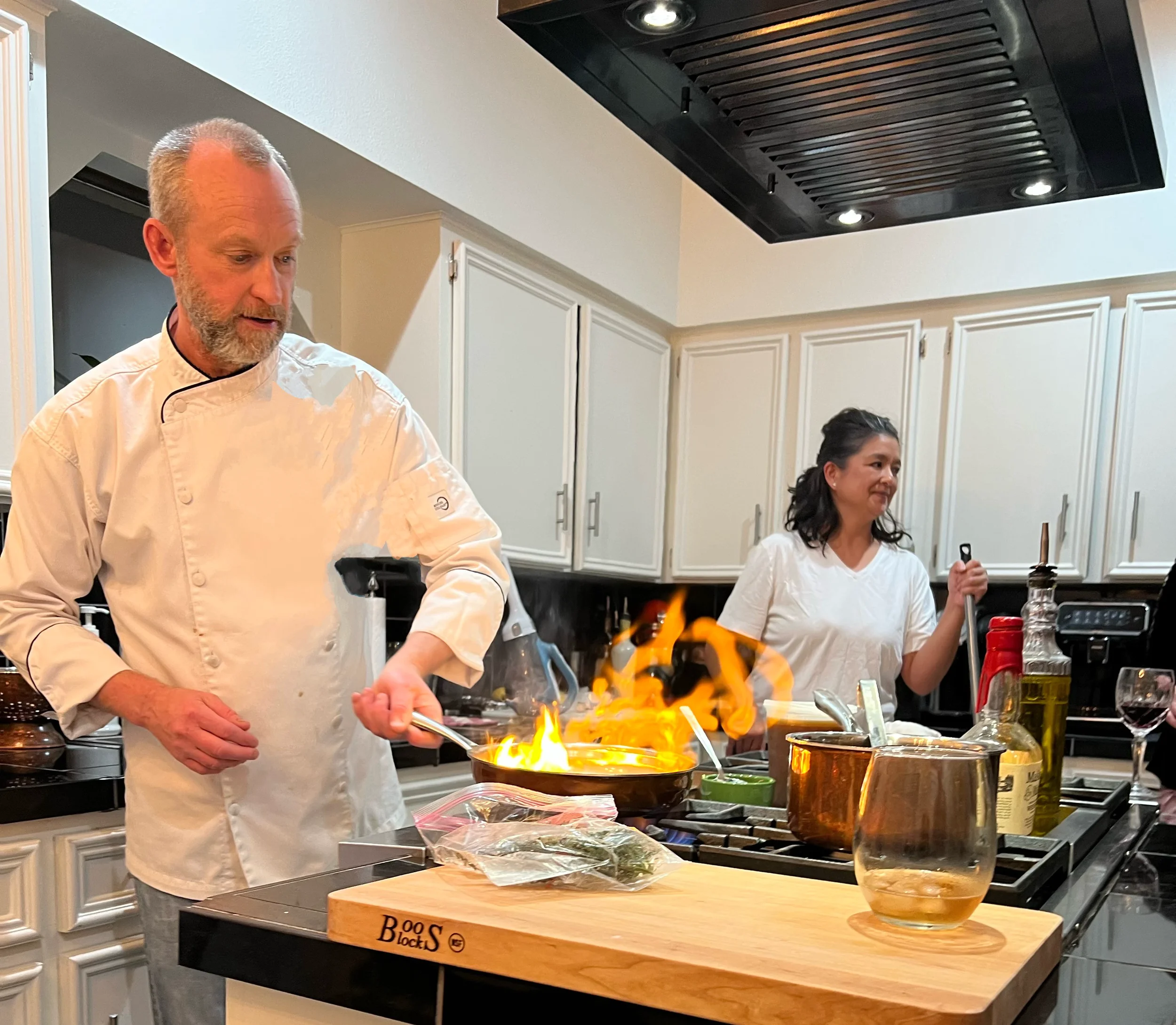 Two people in a kitchen cooking, one man with a white chef's coat has a flame in a pan, and a woman holding a spatula smiles nearby.