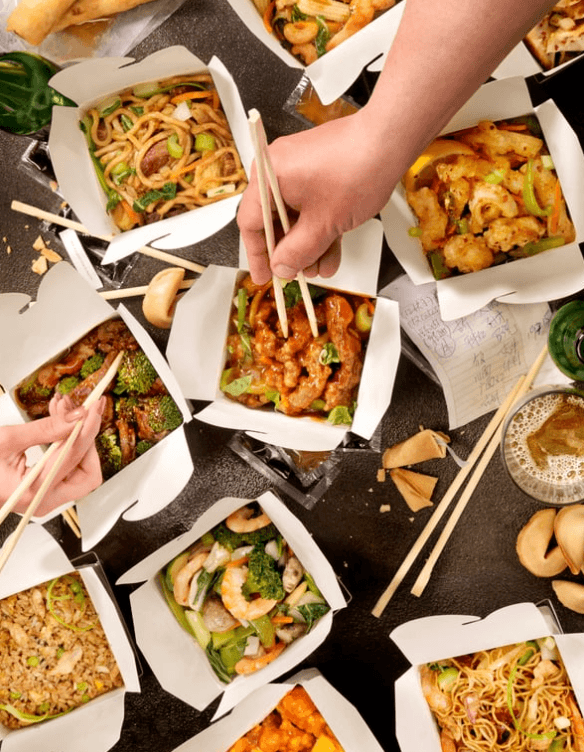 Top-down view of a table with various Chinese takeout boxes filled with noodles, fried rice, vegetables, and chicken, with hands using chopsticks to pick food.