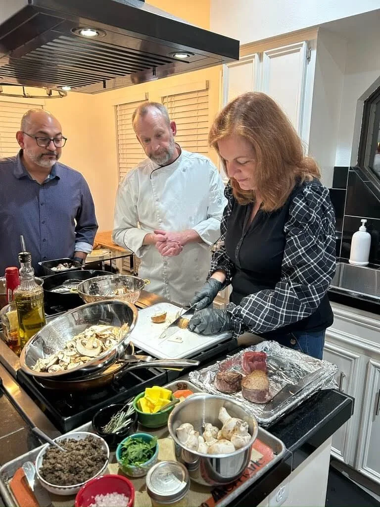 Three people in a kitchen preparing food; a woman is slicing meat, with mushrooms, herbs, and spices on the counter, and a man in a chef's coat and another man in glasses watching.